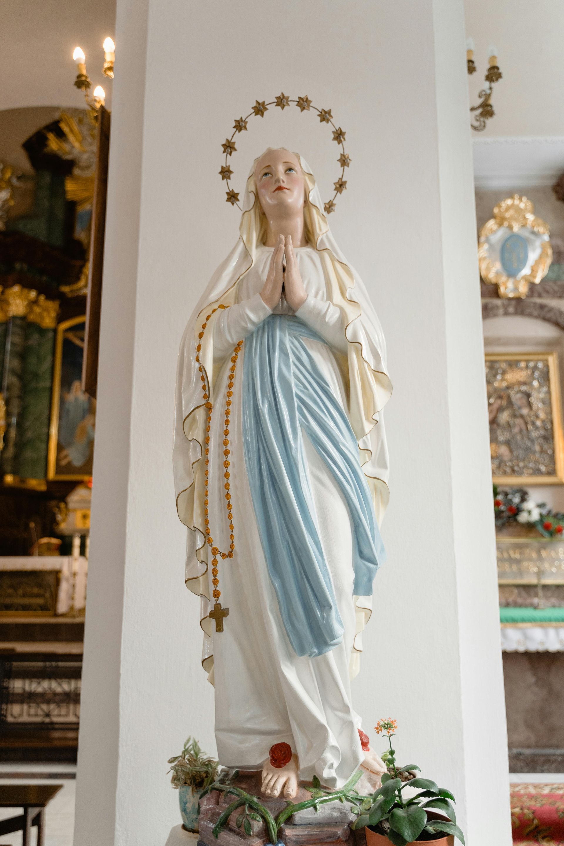 Statue of the Virgin Mary in a white and blue robe, standing in a church alcove with flowers at the base