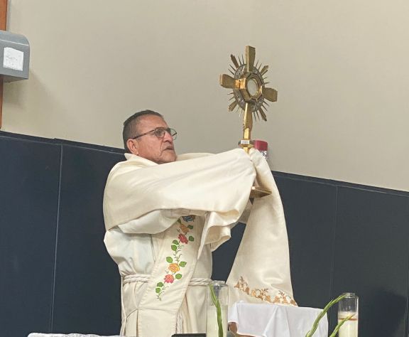 Priest in white vestments holds up a gold monstrance during a church service.