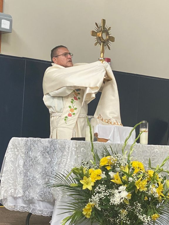 Priest lifting the Eucharist during Mass, standing behind an altar with white cloth and yellow flowers