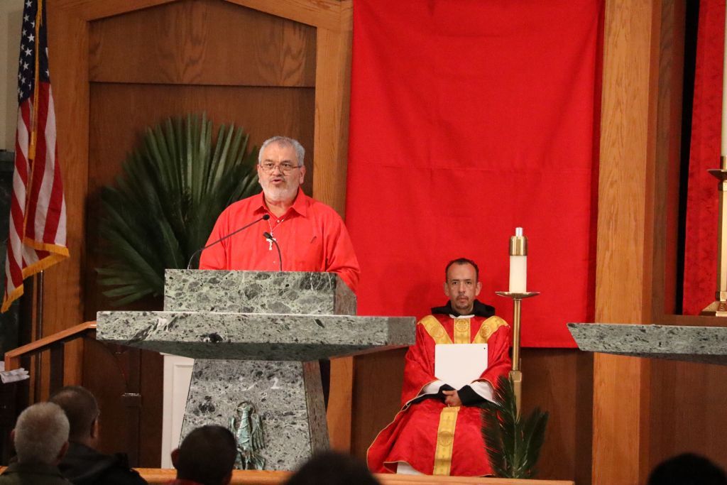 Speaker at stone lectern on stage with seated clergy, red backdrop and American flag