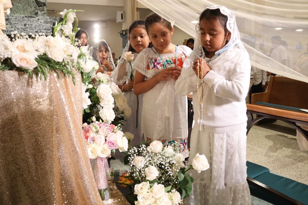 Three women in white stand at a memorial display of flowers and a casket