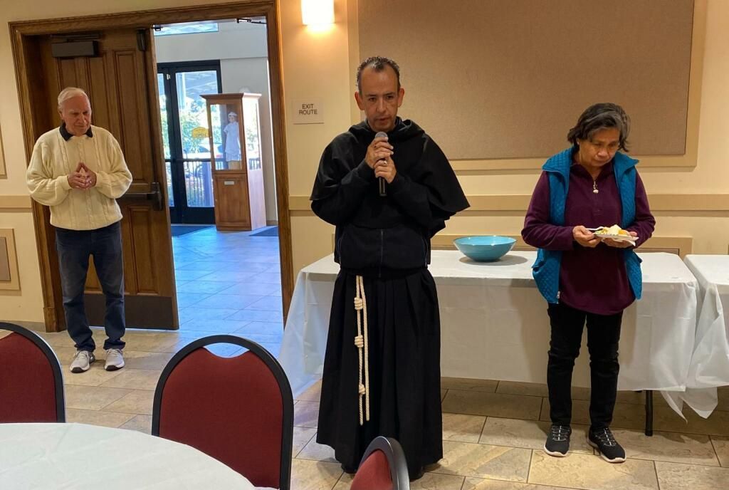 Three people standing in a hall, one man in black speaking into a microphone near a table