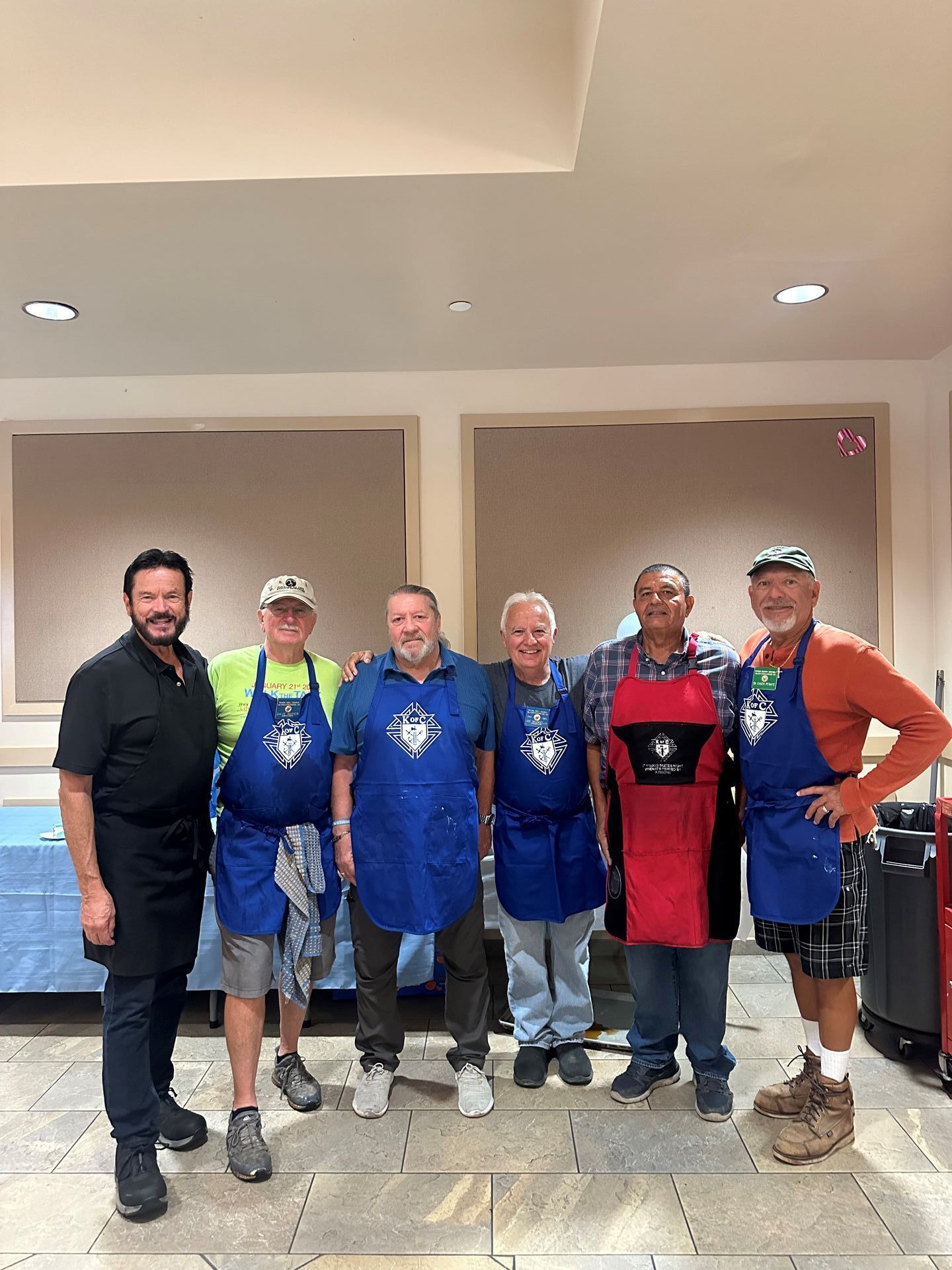 Six men posing indoors, most wearing blue aprons, standing in a lobby-like room.