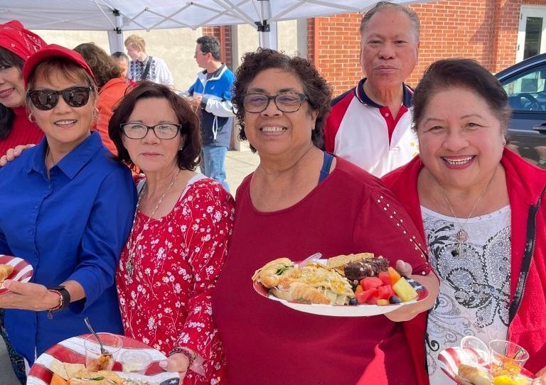 Five people smiling at an outdoor gathering, holding plates of food under a canopy