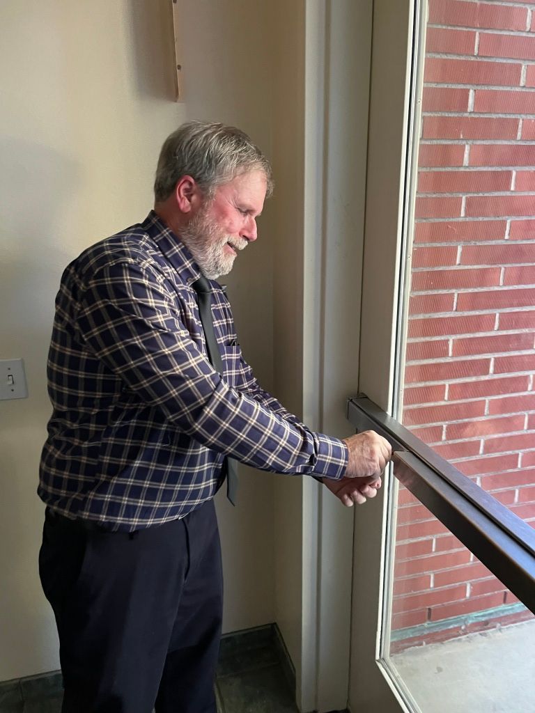 Man standing by a window, holding a railing and looking outside in a plaid shirt.