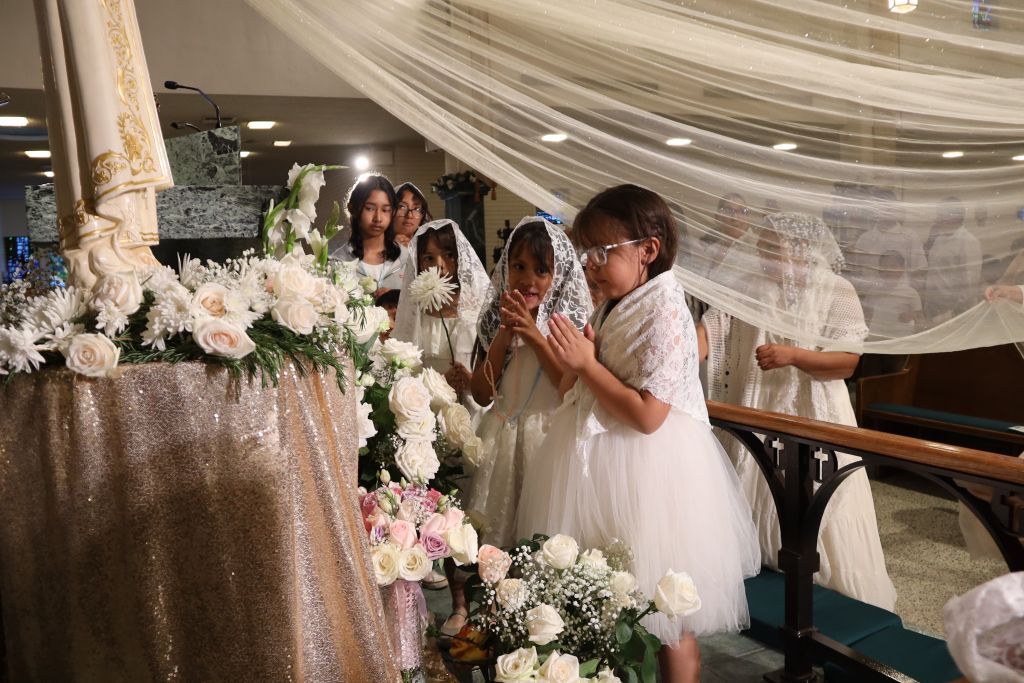 Three people in white at a decorated indoor wedding venue, standing by floral arrangements and draped fabric.