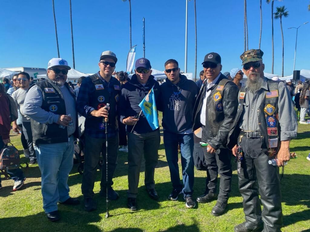 Group of bikers in vests posing outdoors on grass, holding flags under a clear blue sky.