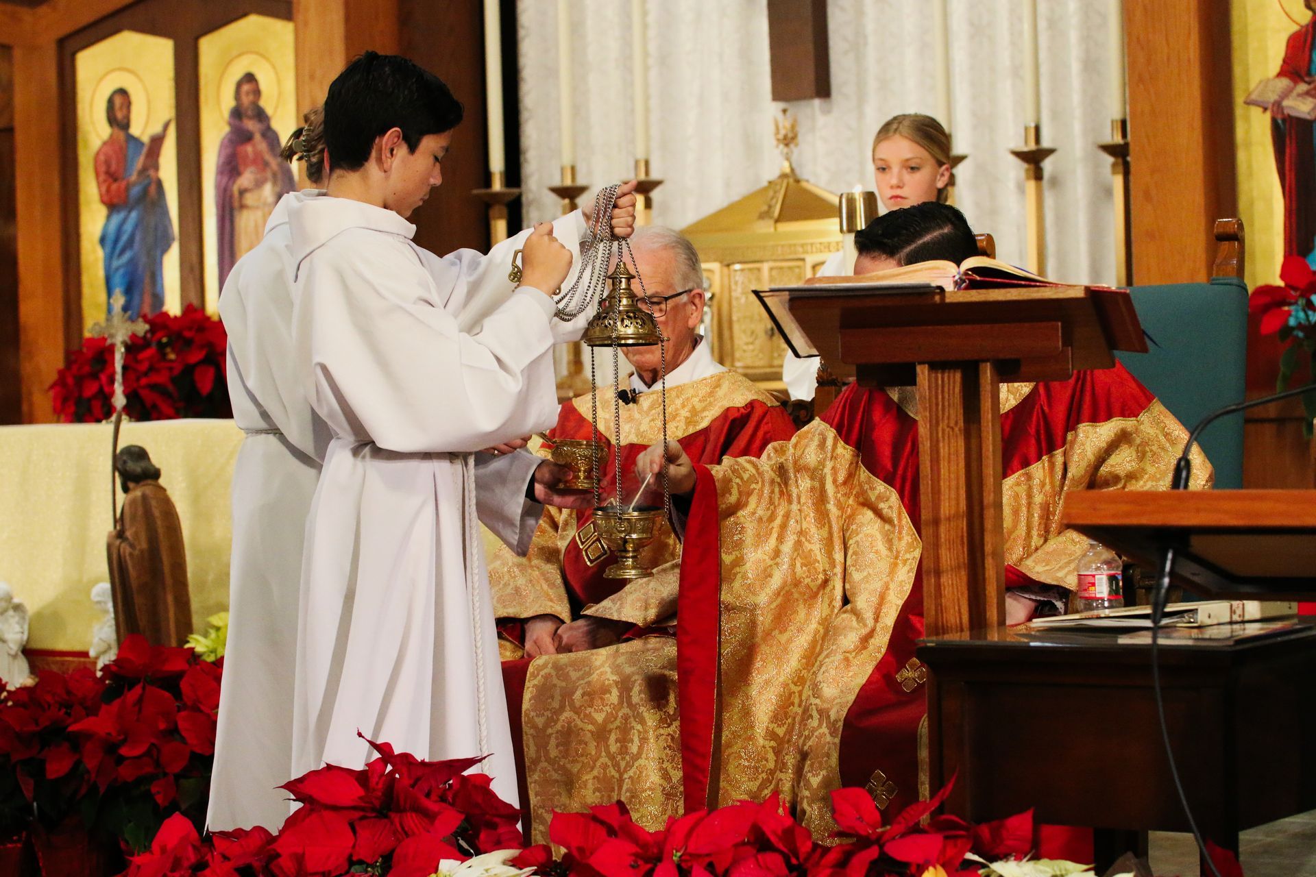 Priest being crowned in a church ceremony, with altar server and red flowers in the foreground