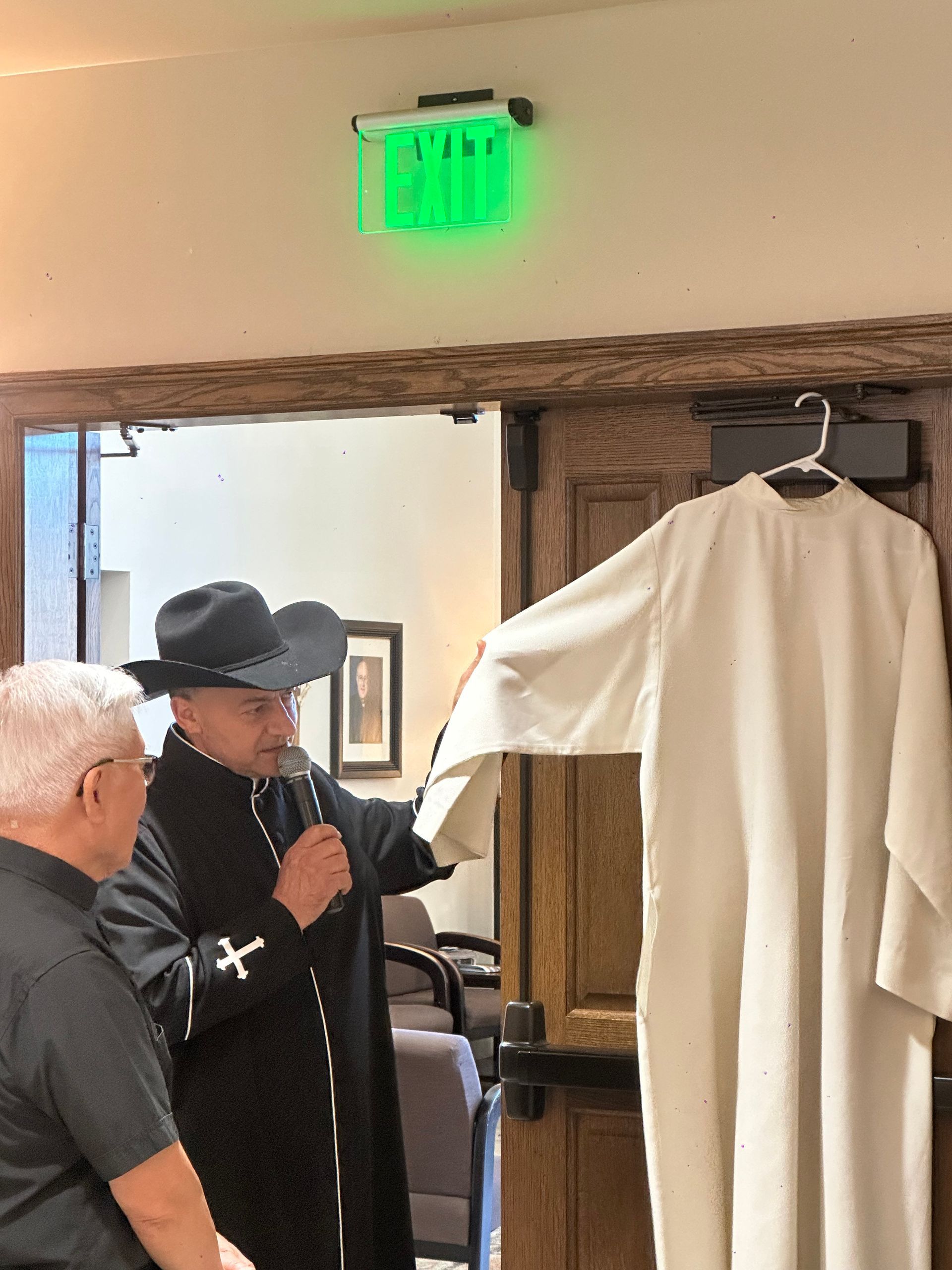 Man in black hat speaks into microphone beside a white garment hanging in a doorway, under a green exit sign