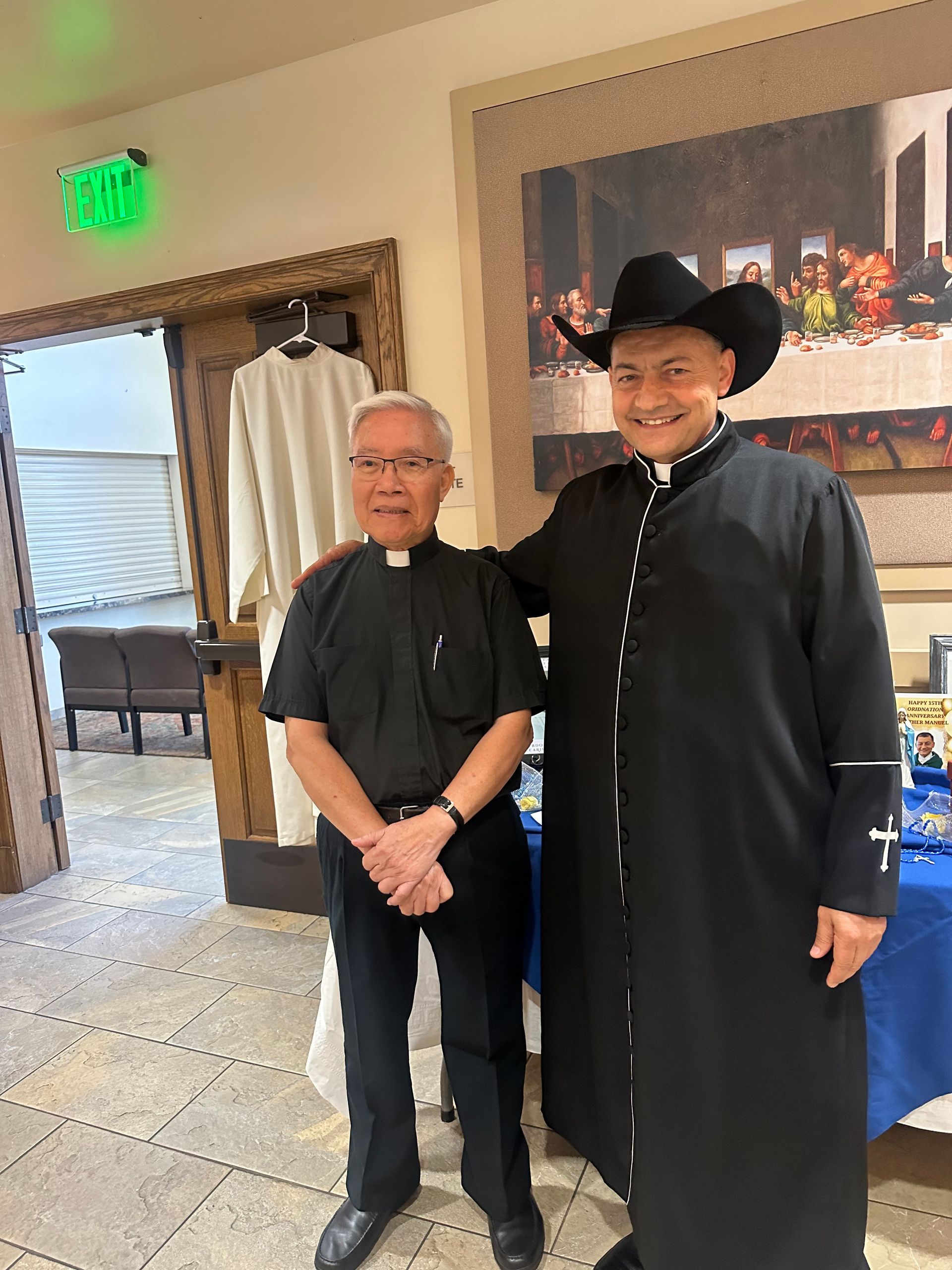 Two people posing indoors, one in a black suit and hat, beside a display table and framed wall art.