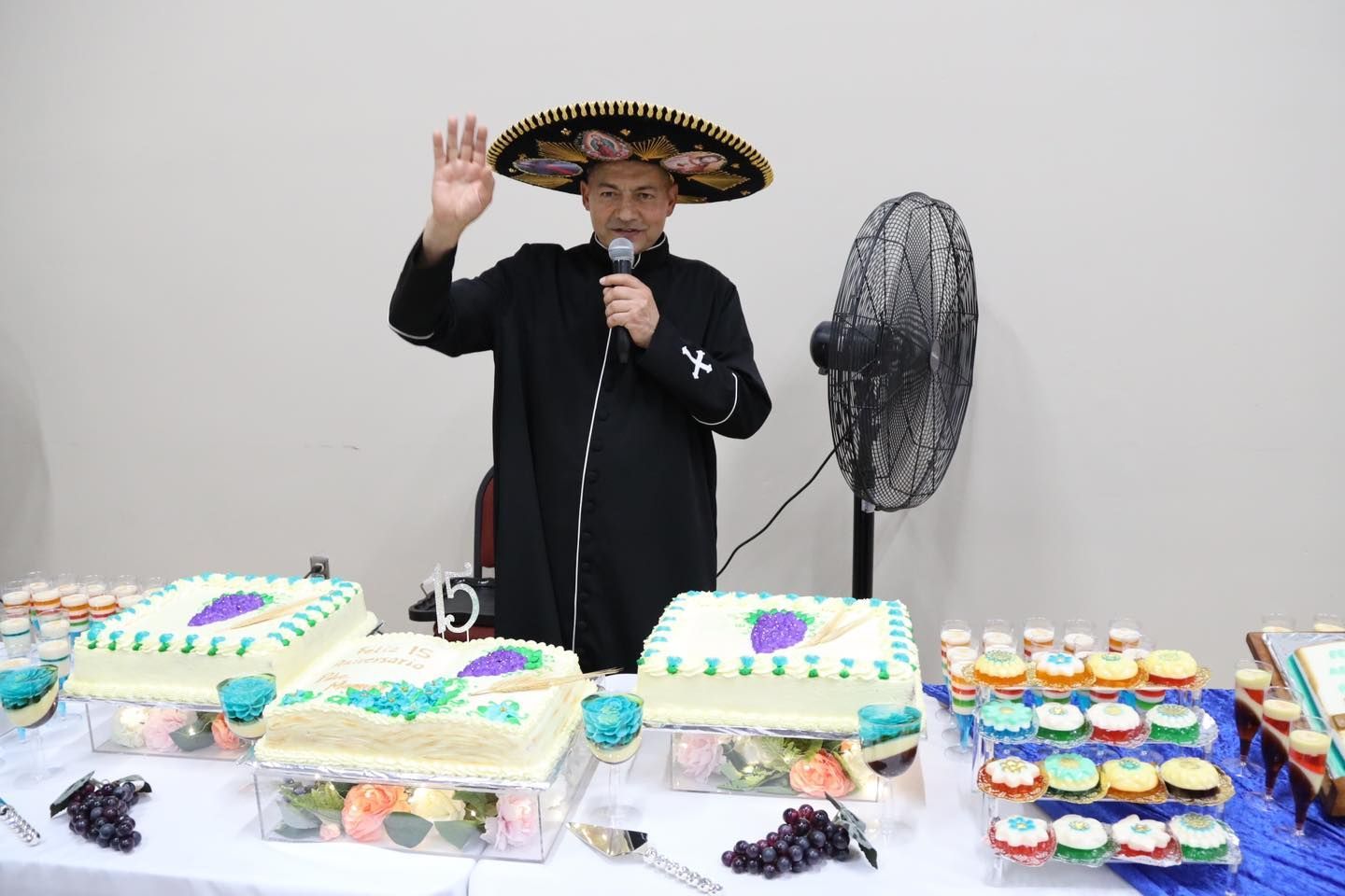 Man in a sombrero speaking at a dessert table with cakes, cupcakes, and a standing fan behind him
