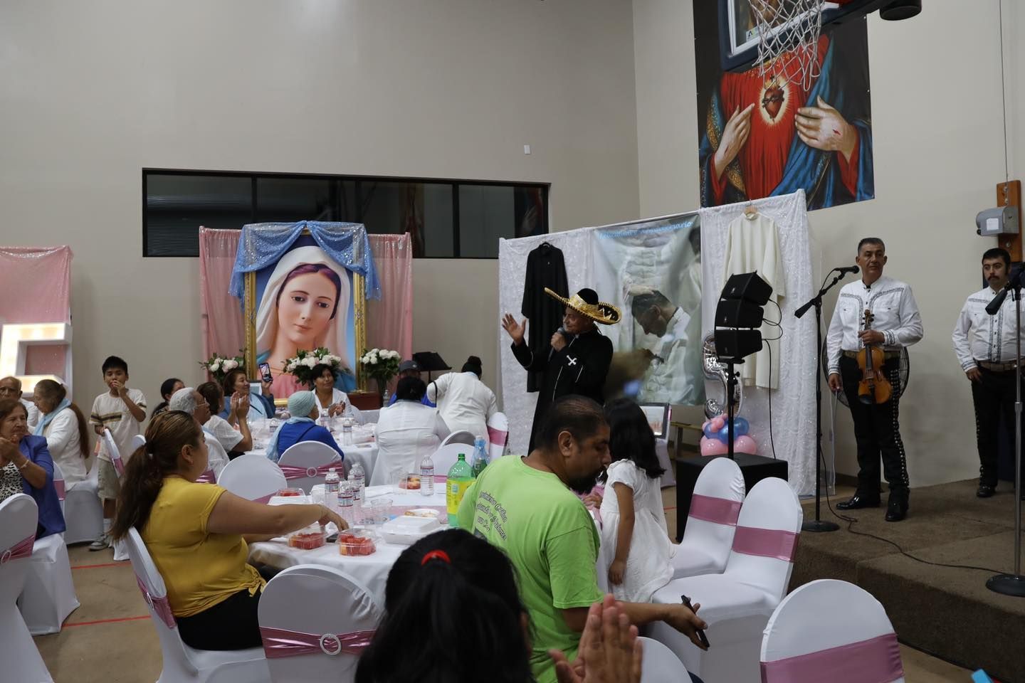 People seated at decorated tables in a hall, with a portrait, white drapes, and musicians performing nearby.