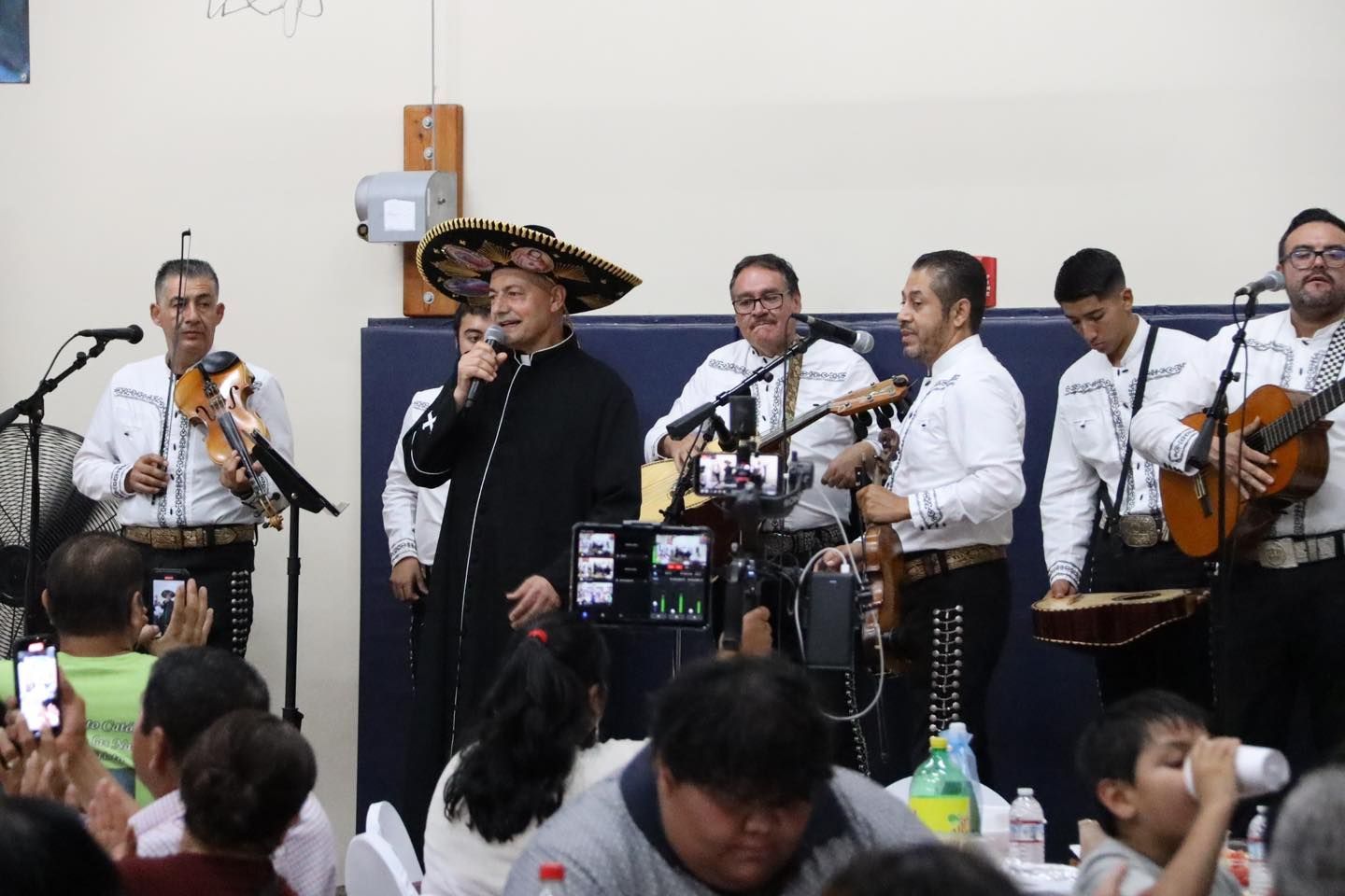 Mariachi band performing indoors with singers and string players in white shirts and sombrero, audience seated nearby