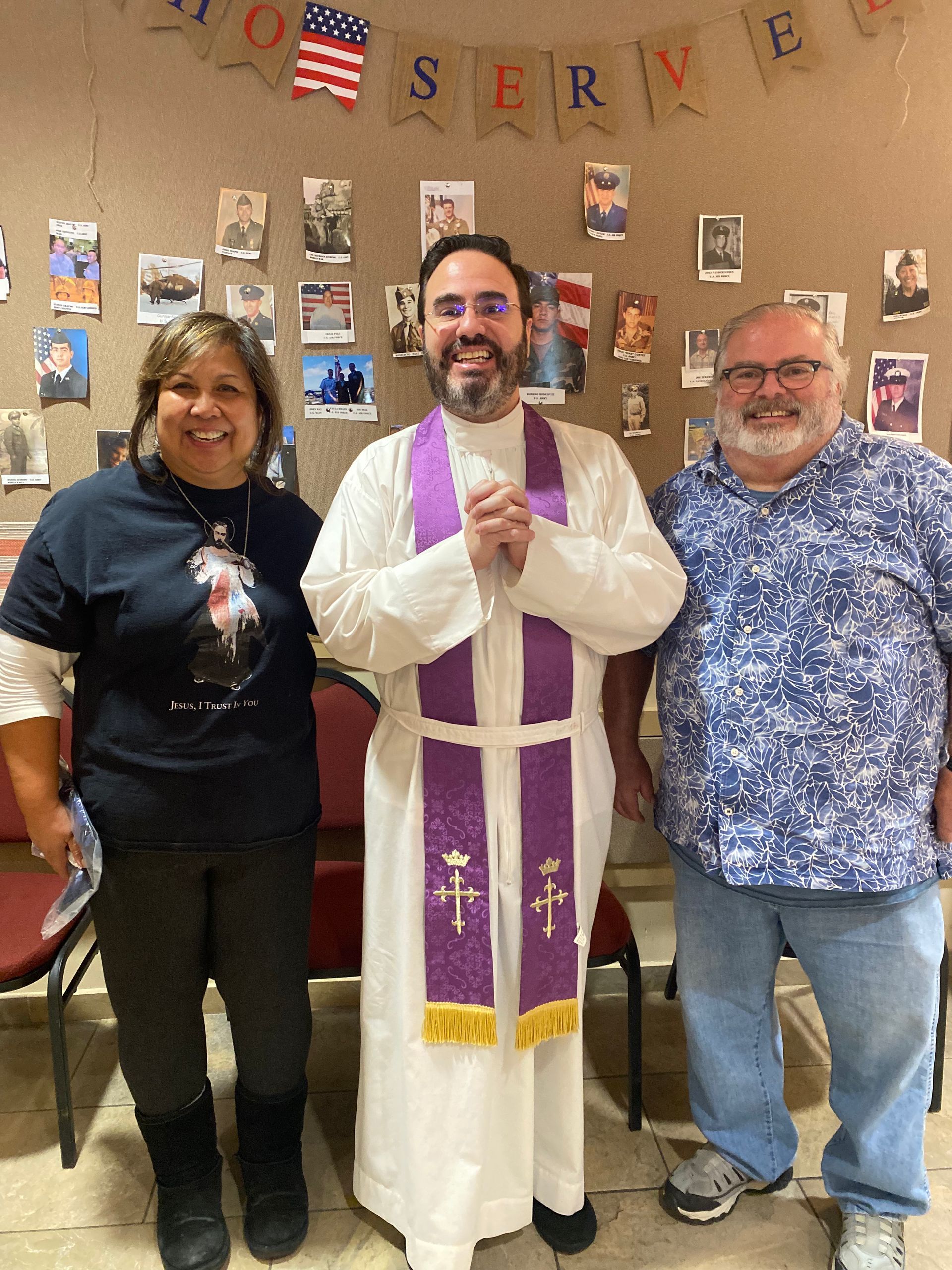 Three smiling people standing indoors, with a priest in white vestments and purple stole centered between two others