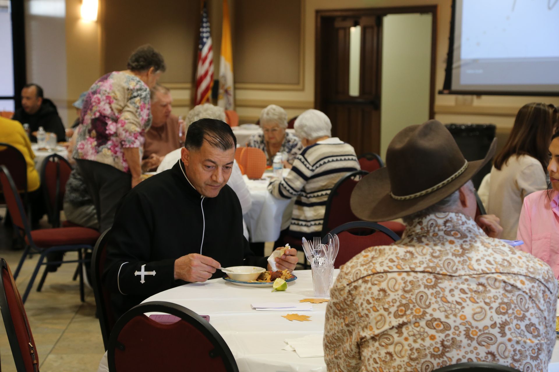 People seated at round tables in a community hall during a gathering, with a projected screen in front.