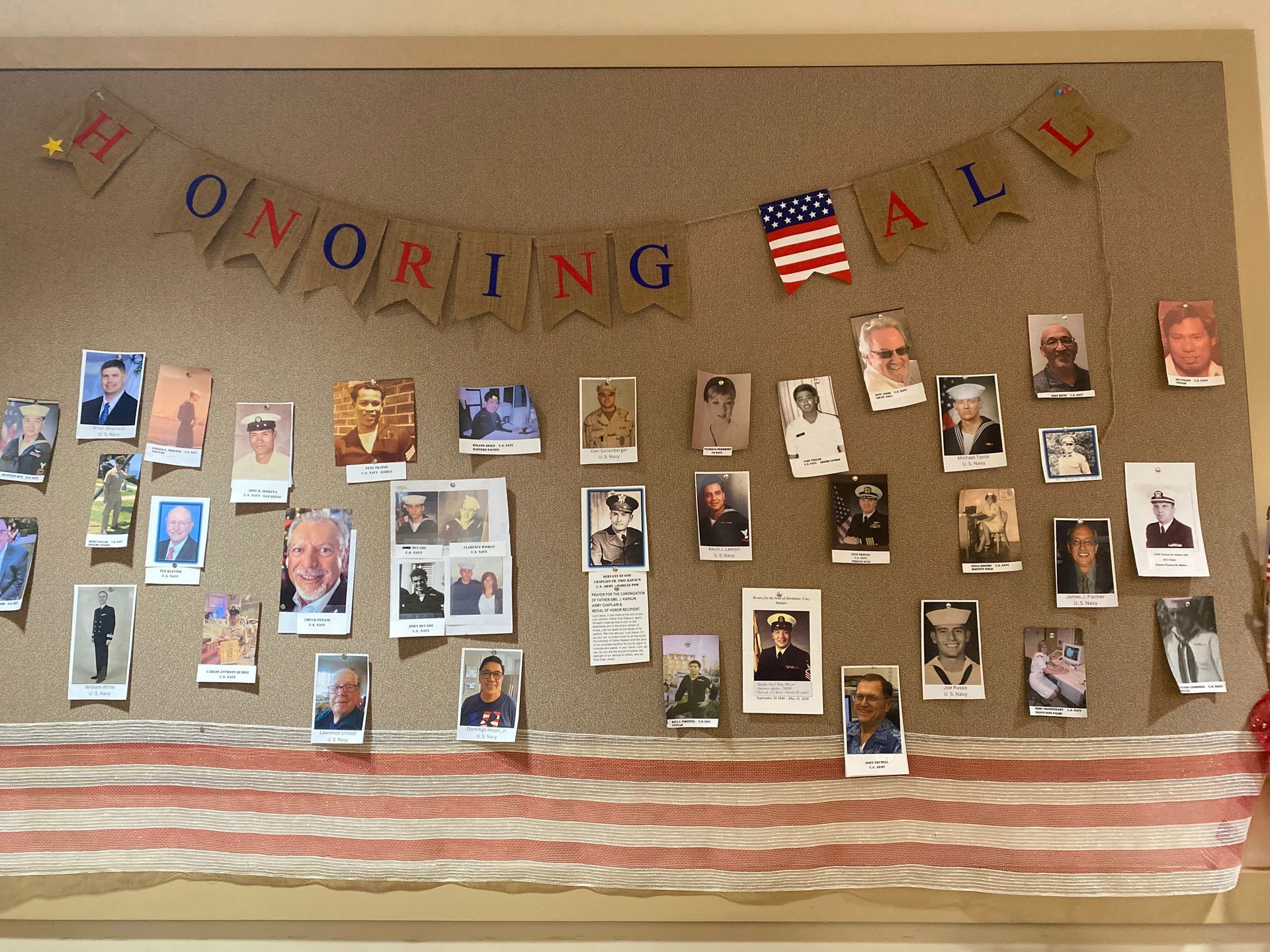 Memorial bulletin board with many photos and “Honoring All” banner above a striped cloth drape.
