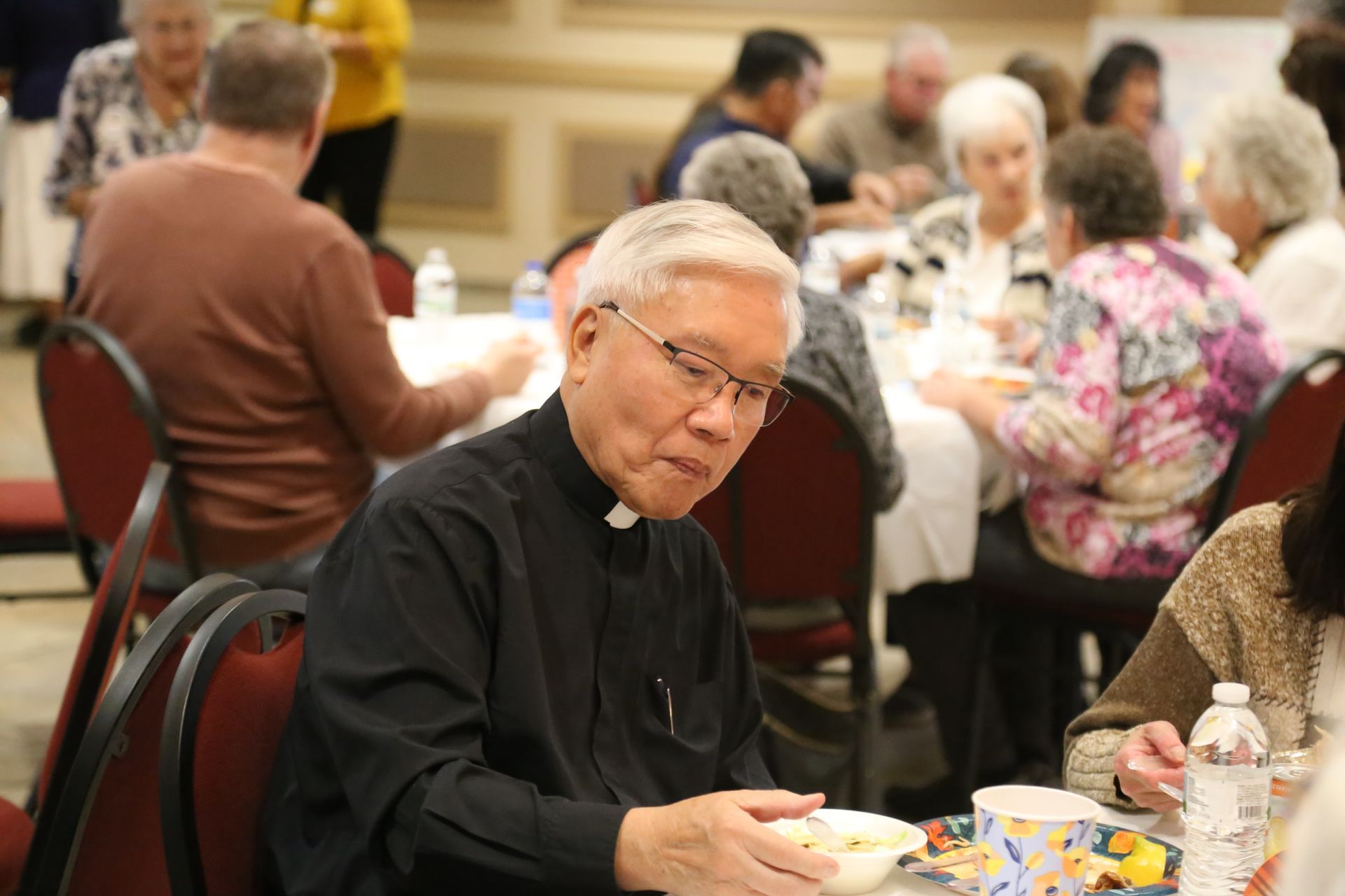 Man in black shirt eating at a banquet table, looking toward the camera, with other diners in the background.