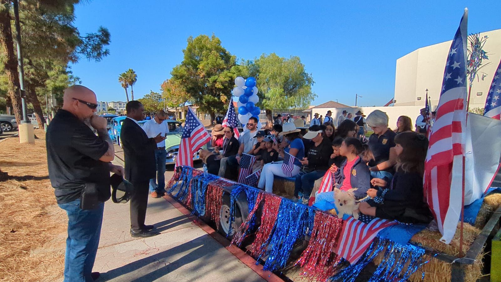 Outdoor patriotic gathering with a speaker addressing a crowd behind red, white, and blue decorations