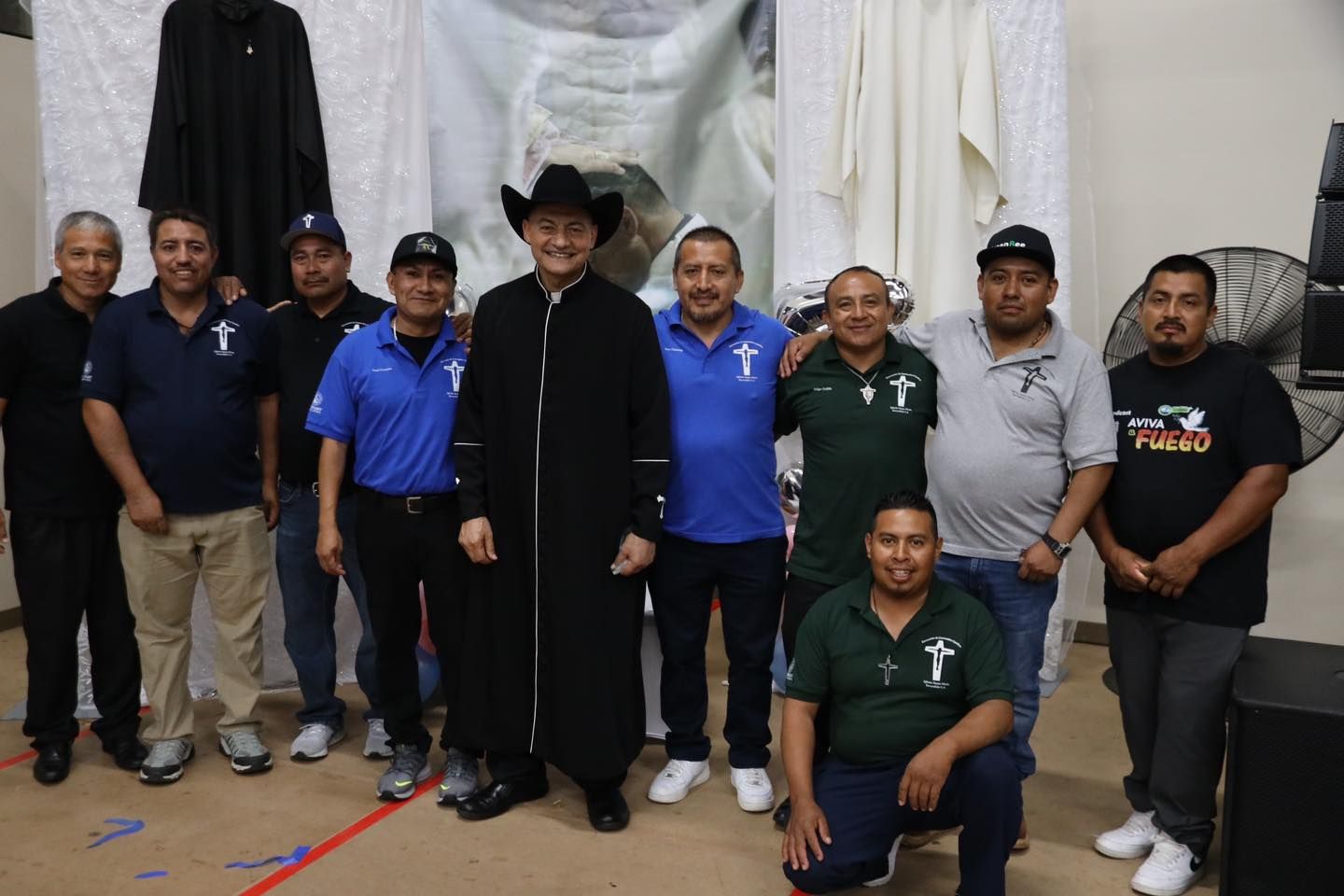 Group of people posing indoors, with one person in a black hat and robe centered under hanging garments.