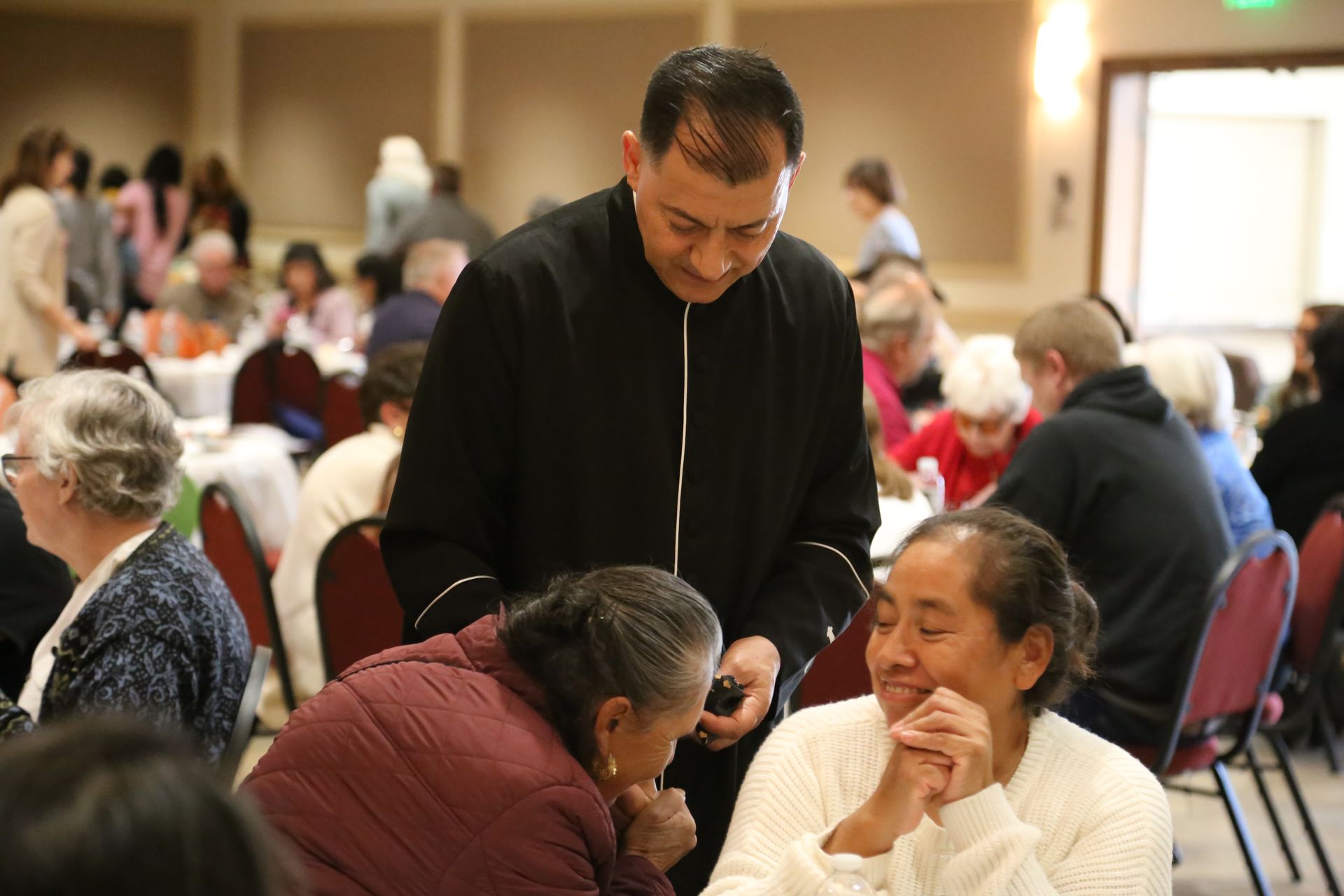 People chatting at a community gathering in a banquet hall, with a man leaning over to speak to seated guests.