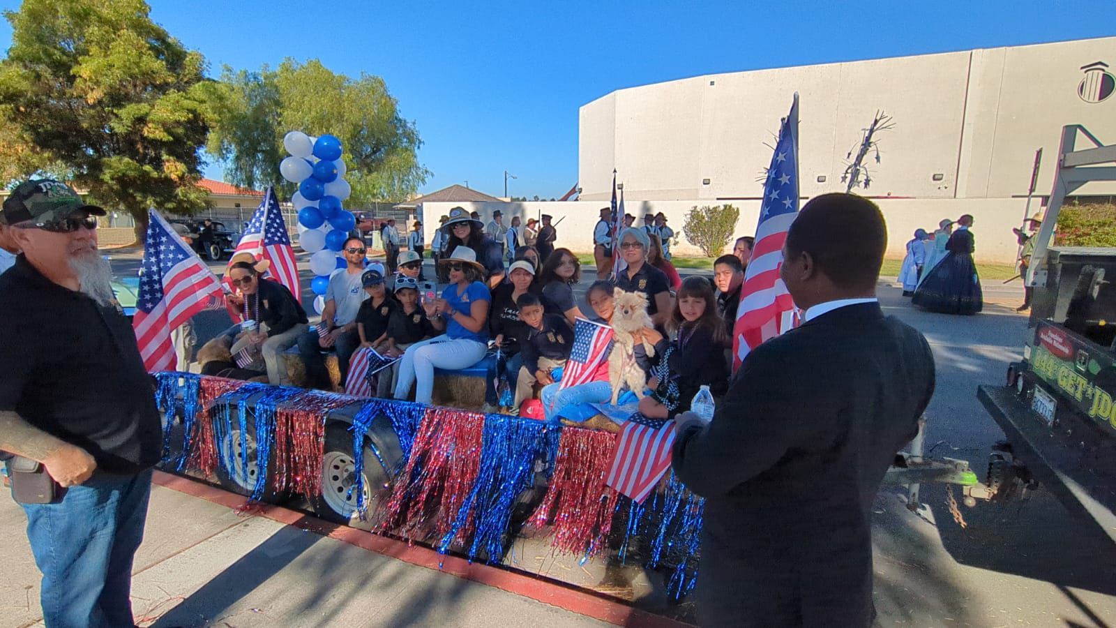 Outdoor patriotic rally with people seated at a decorated table, American flags, and blue balloons near a building.