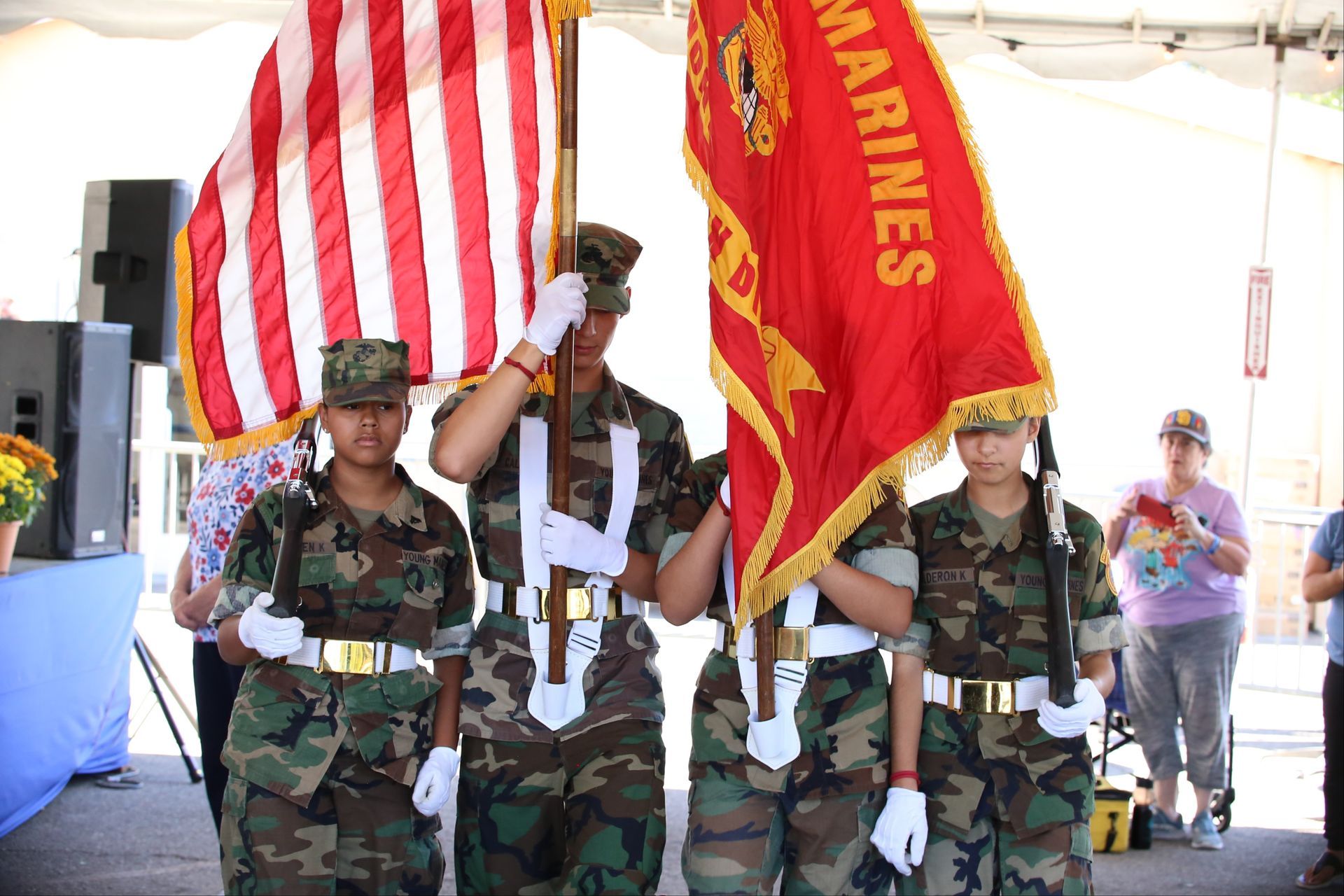 Four Marines in dress uniforms hold flags at a ceremonial event outdoors.