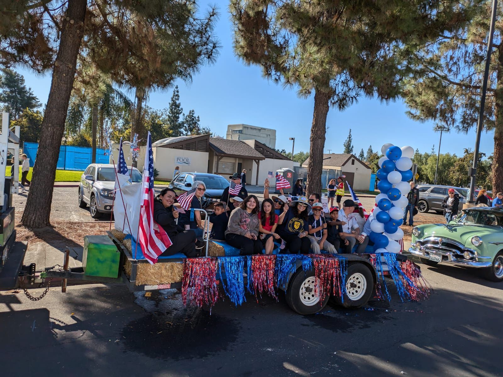 Decorated parade float with seated people, blue-white balloon arch, and patriotic flags in a tree-lined street