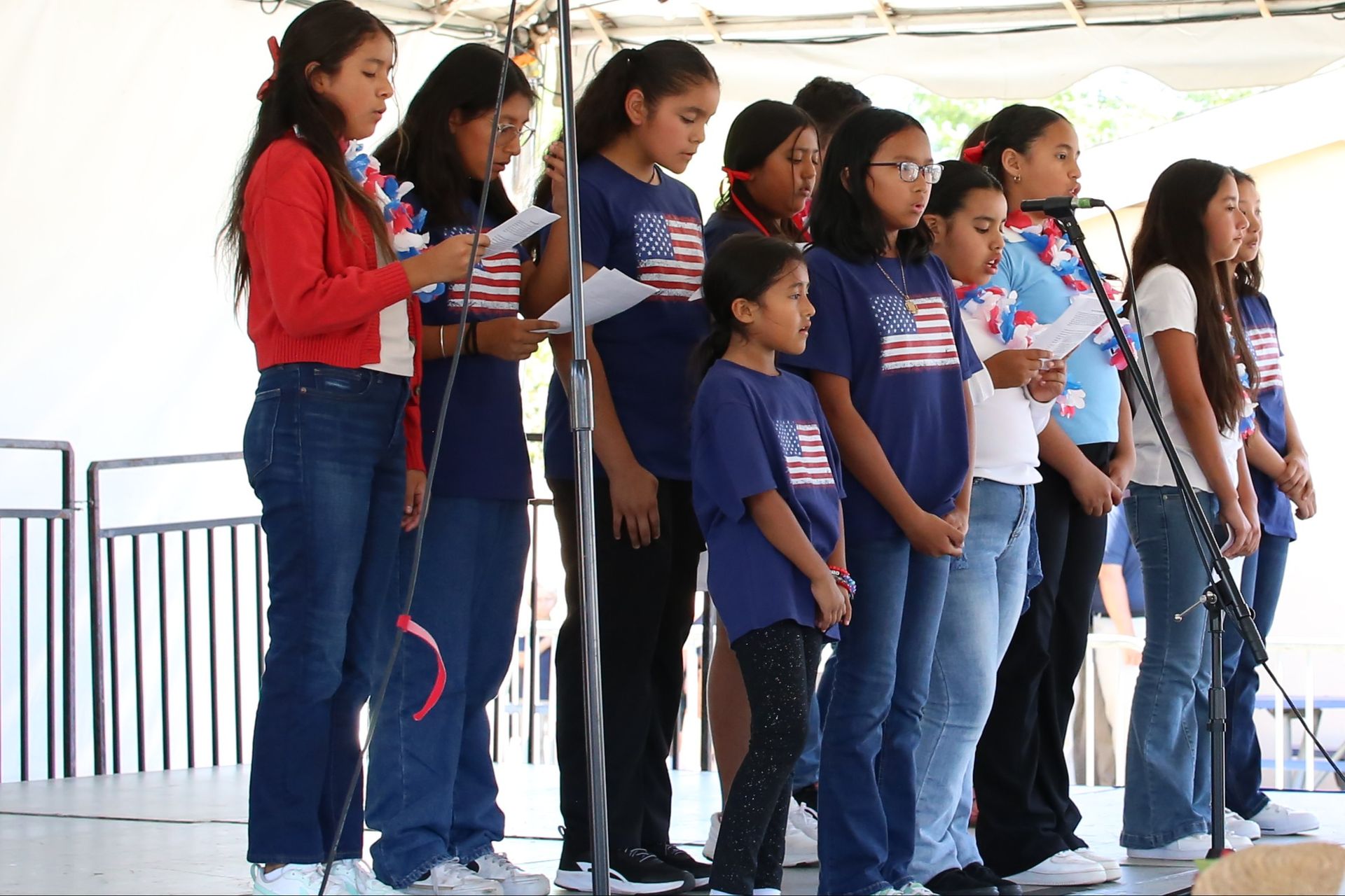 Group of children in red, white, and blue shirts singing on a stage with microphones