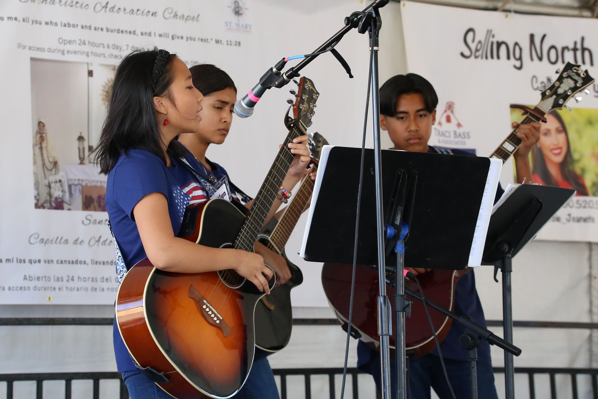 Three musicians perform with string instruments and microphones on stage at an indoor event.