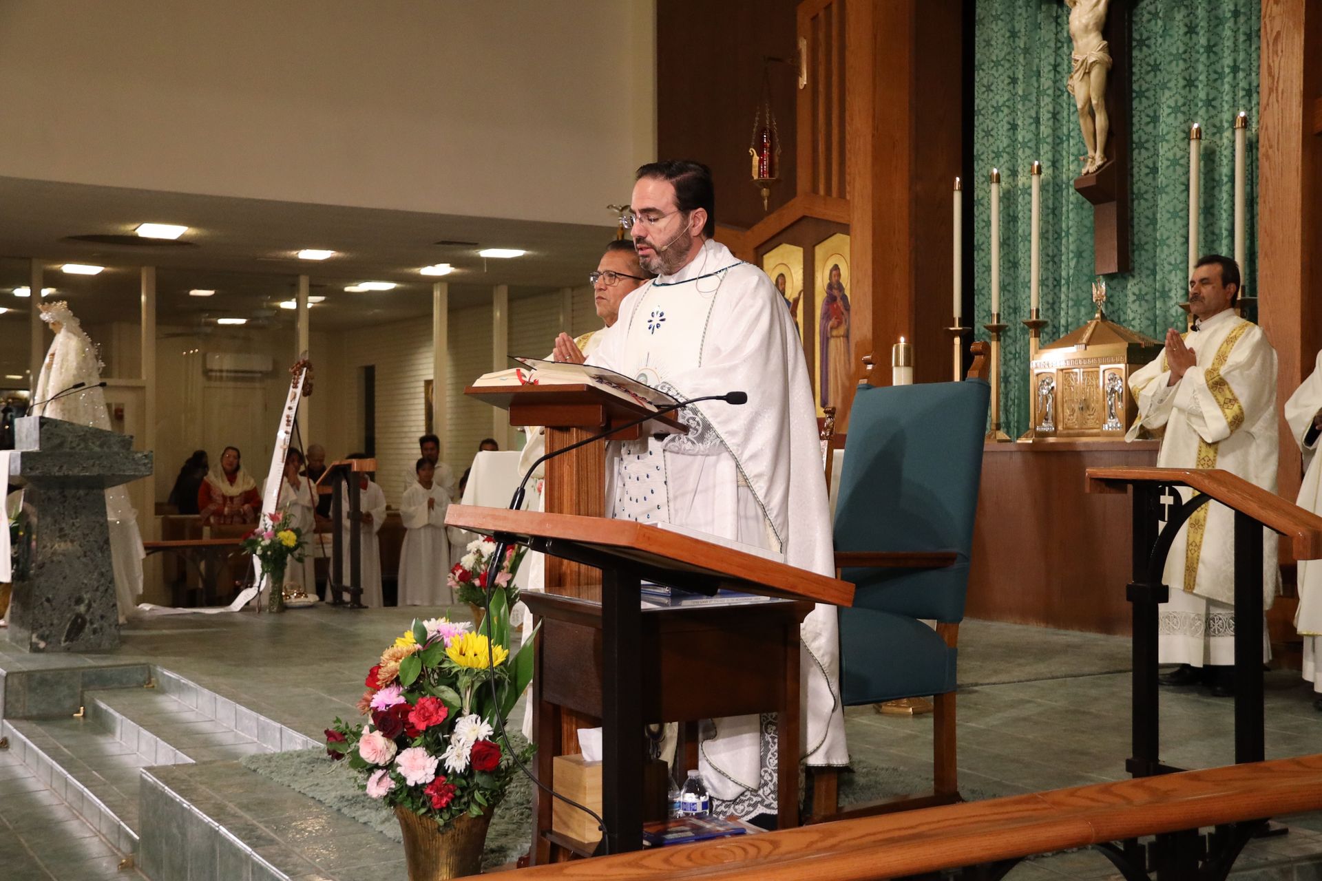 Priest speaking at church lectern during Mass, with altar servers and flowers nearby.