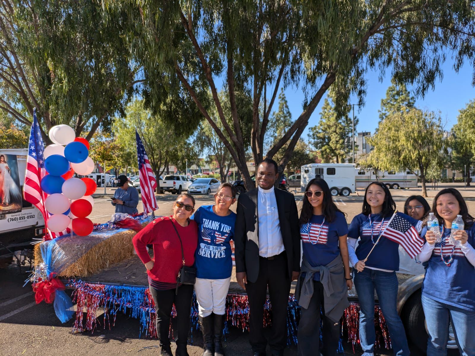 Group posing outdoors by a red-white-blue decorated car, holding small American flags under trees.