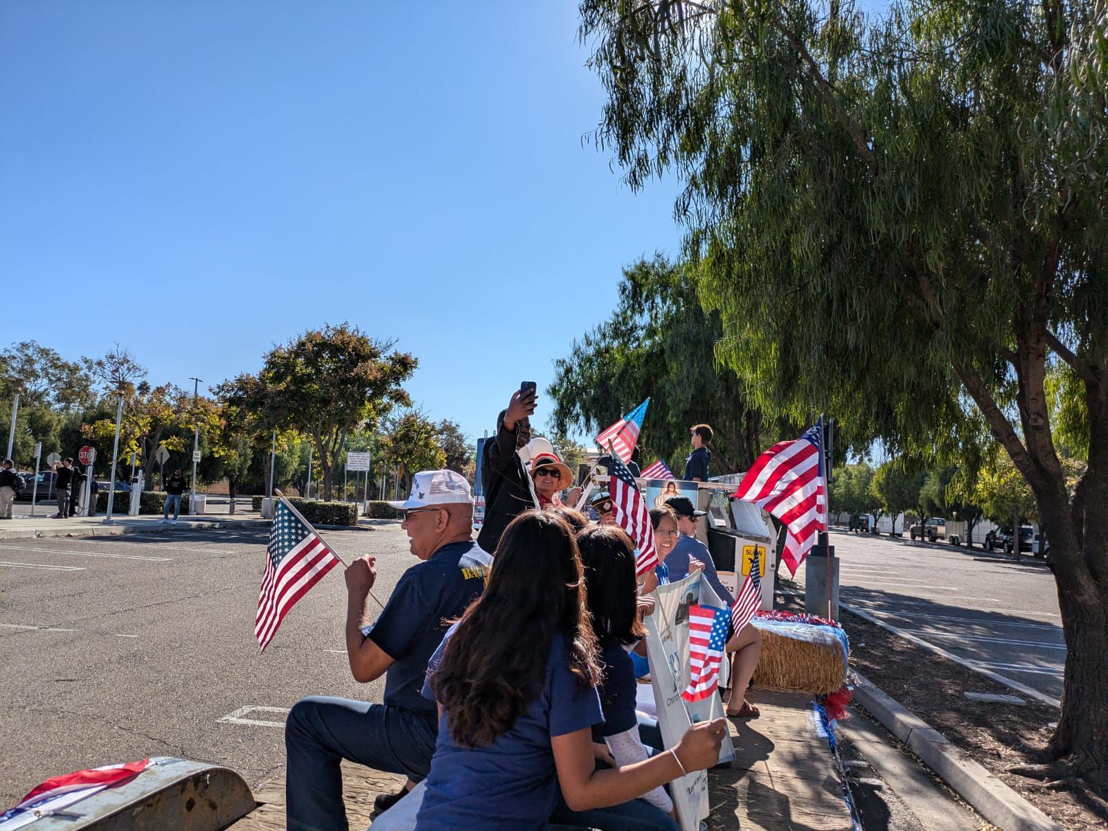 People holding small American flags at an outdoor gathering under trees on a sunny day