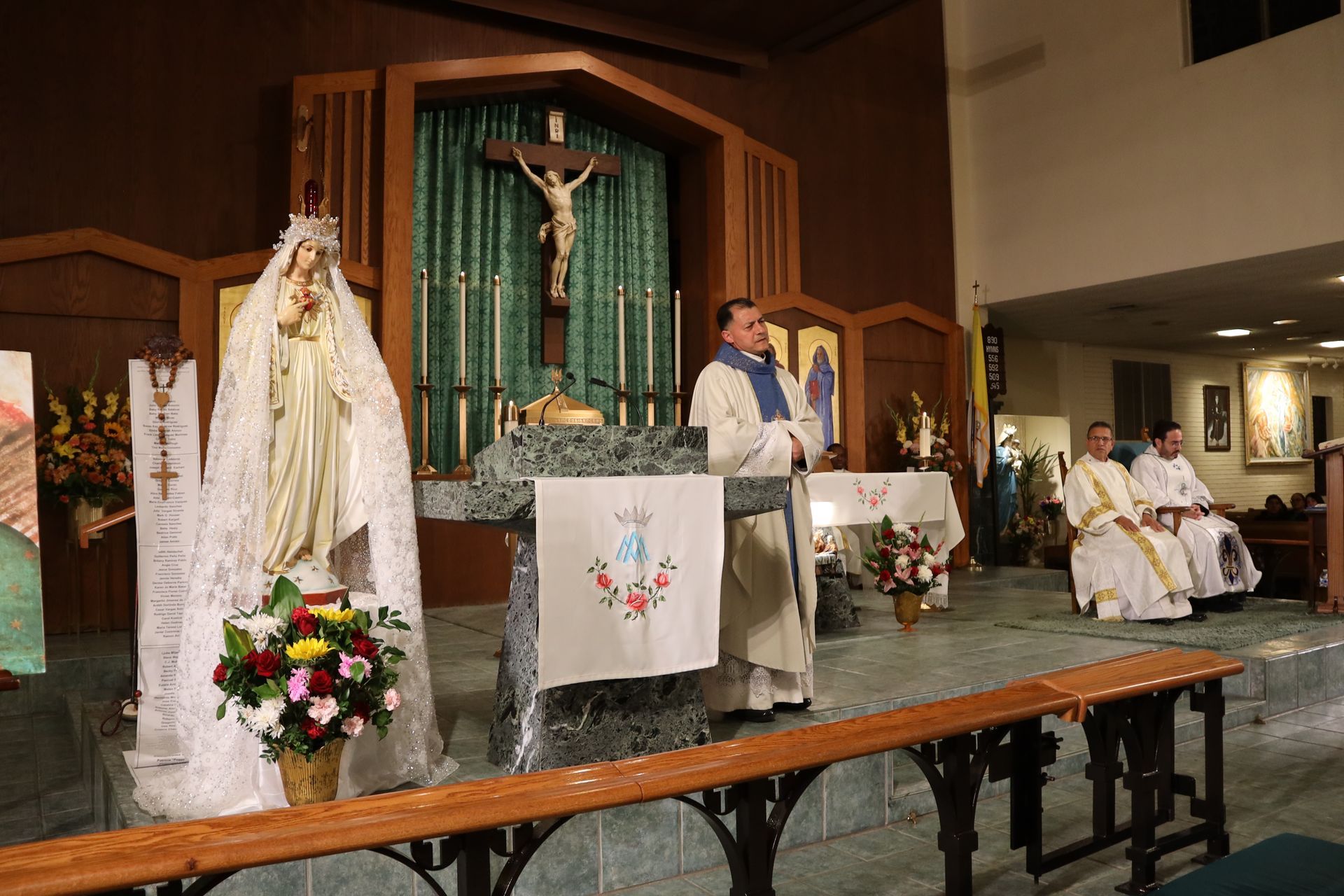 Catholic church altar with crucifix, floral arrangements, and clergy seated during a service