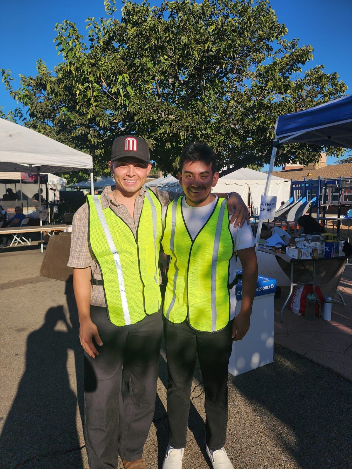 Two people in neon safety vests standing outdoors at a sunny event booth.