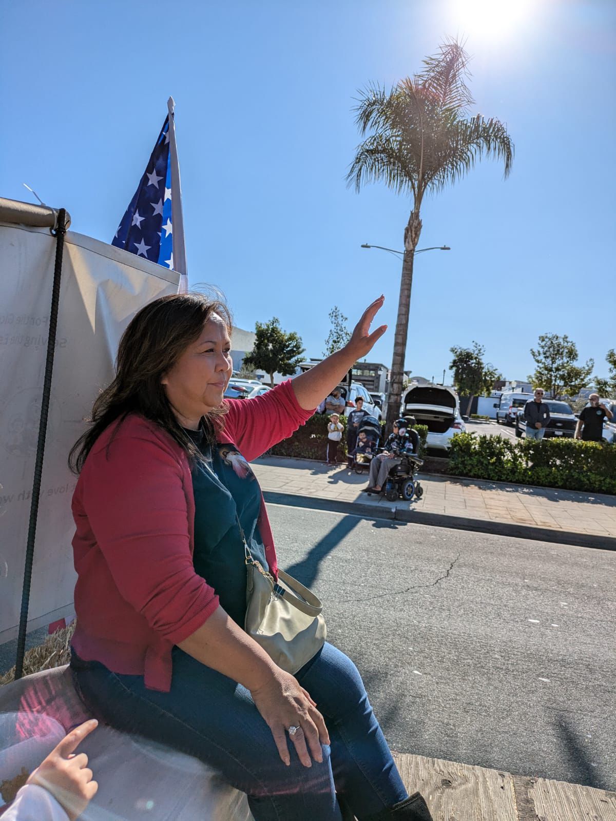 Woman in pink jacket gesturing outdoors beside a tent and flag on a sunny street