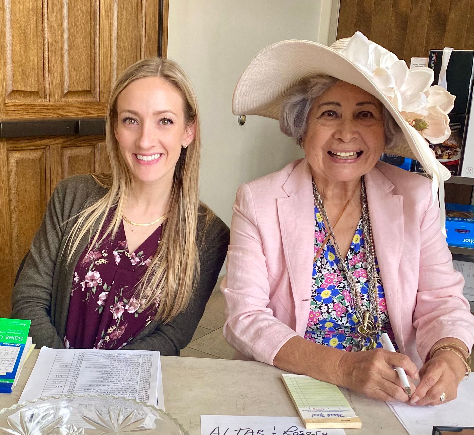 Two women smiling at a table, one wearing a pink blazer and wide-brim hat, in a kitchen.