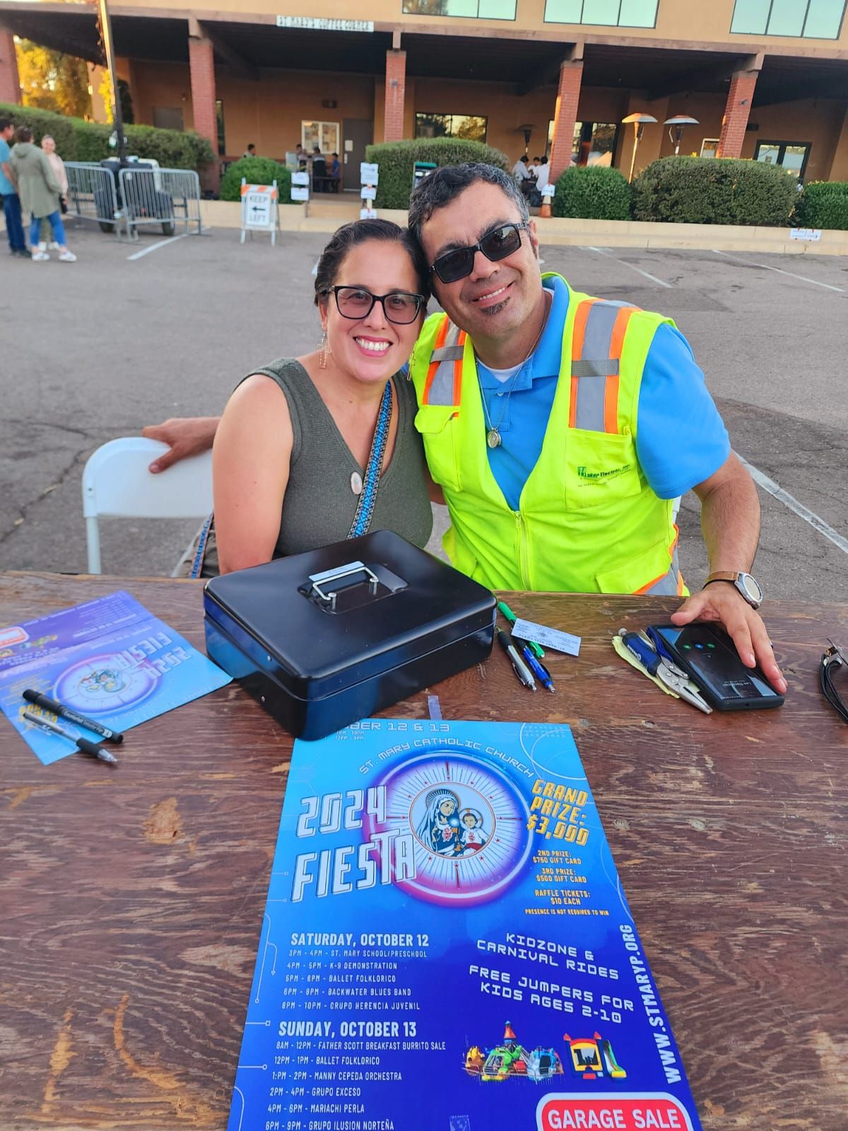 Two smiling people at a table with Fiesta flyers and a laptop at an outdoor event.