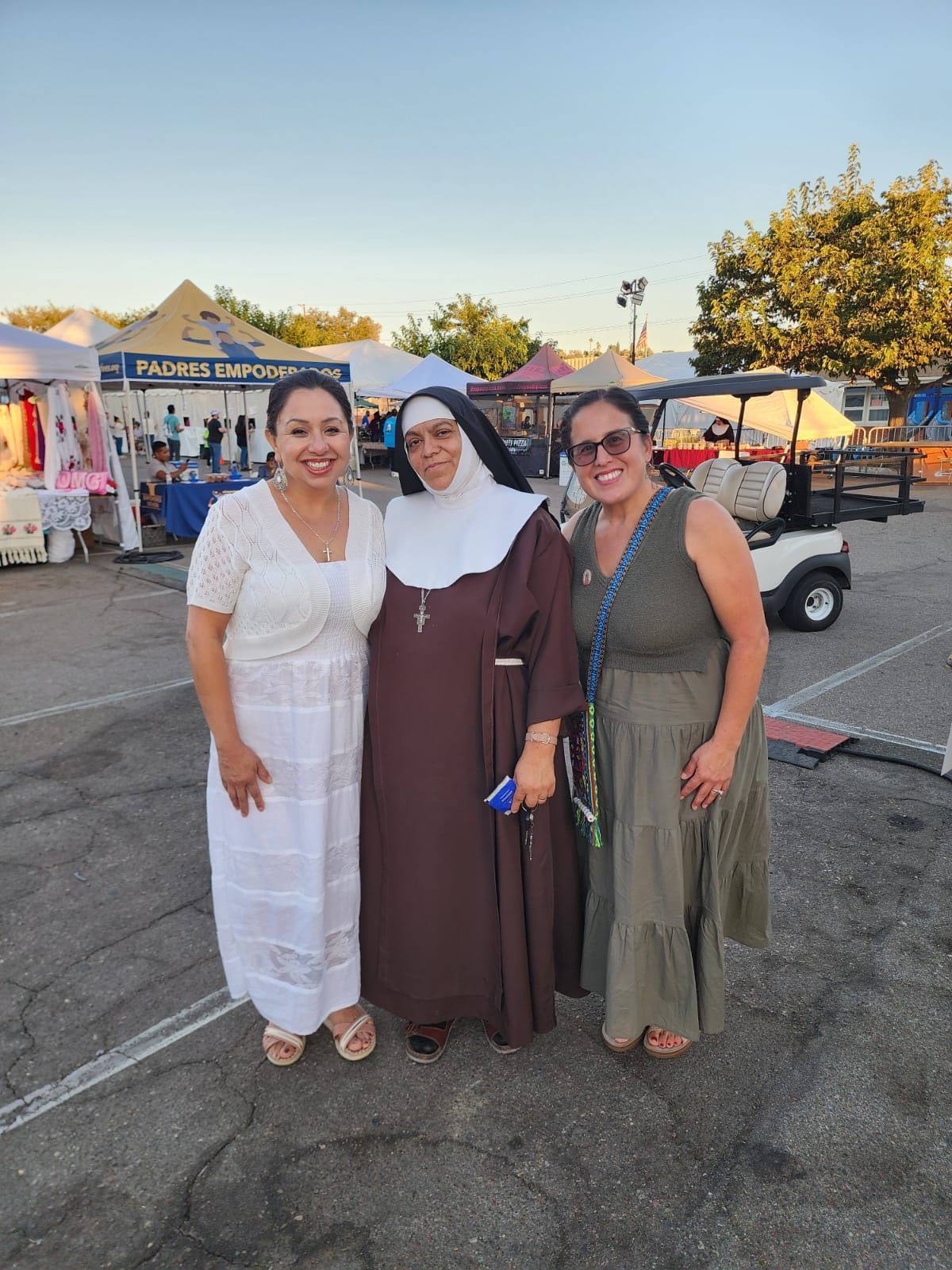 Three women posing together at an outdoor market at dusk, with stalls and a golf cart in the background.