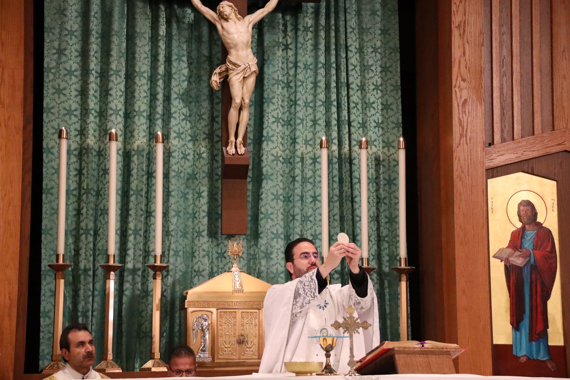 Priest elevates a chalice at the altar beneath a crucifix, flanked by candles and a religious icon.