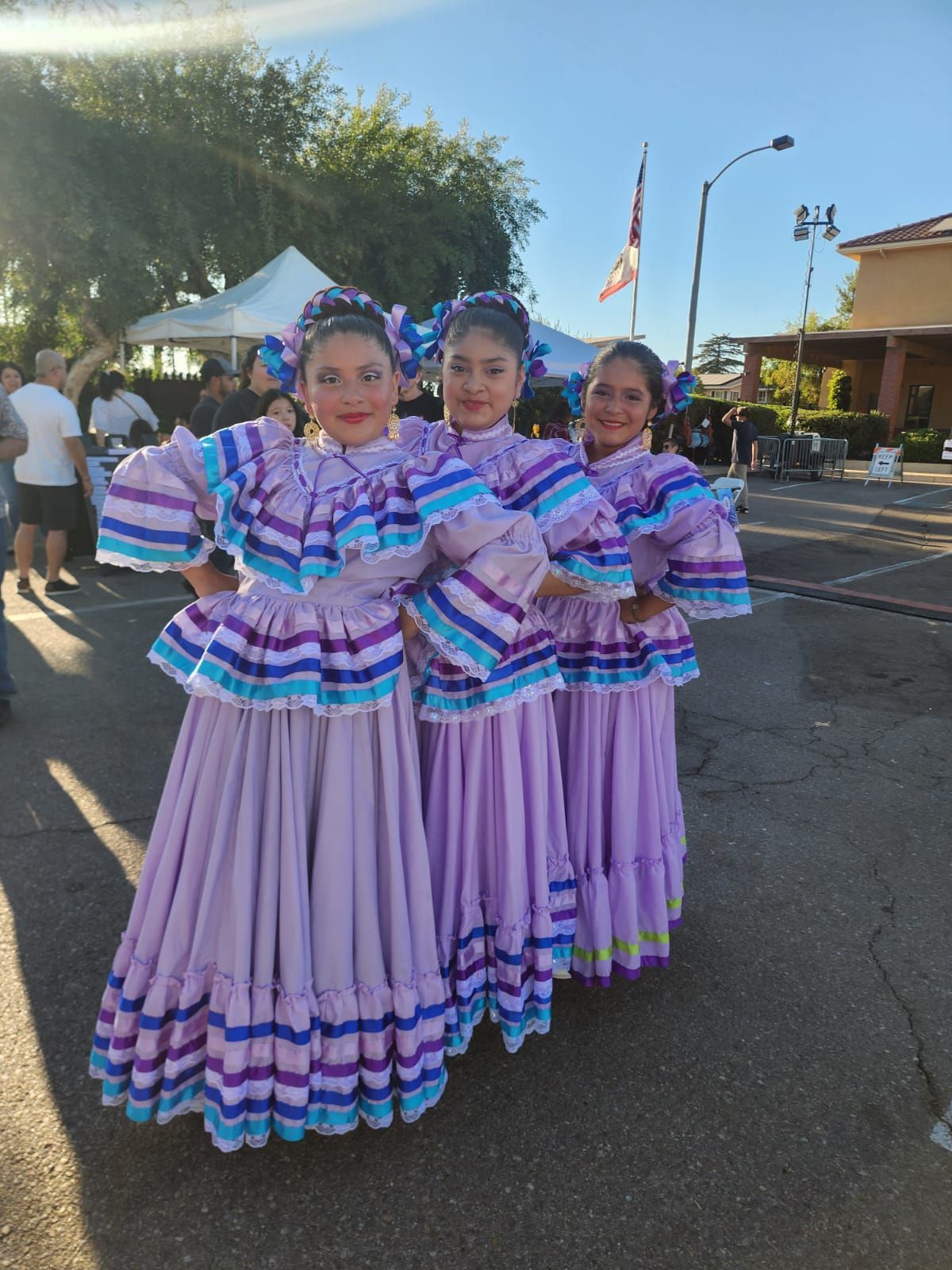 Three people in matching lavender folkloric dresses posing outdoors at a parade