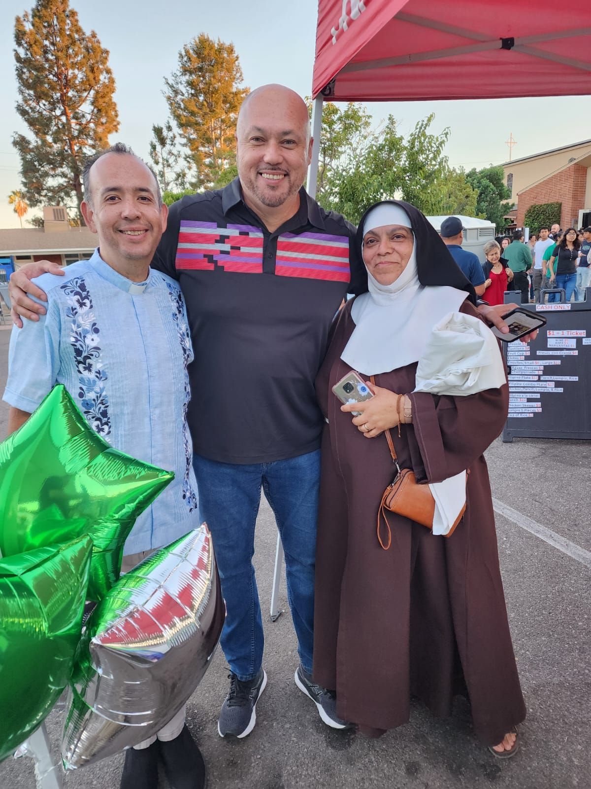 Three people posing outdoors at a festive event, with balloons and a red canopy in the background.