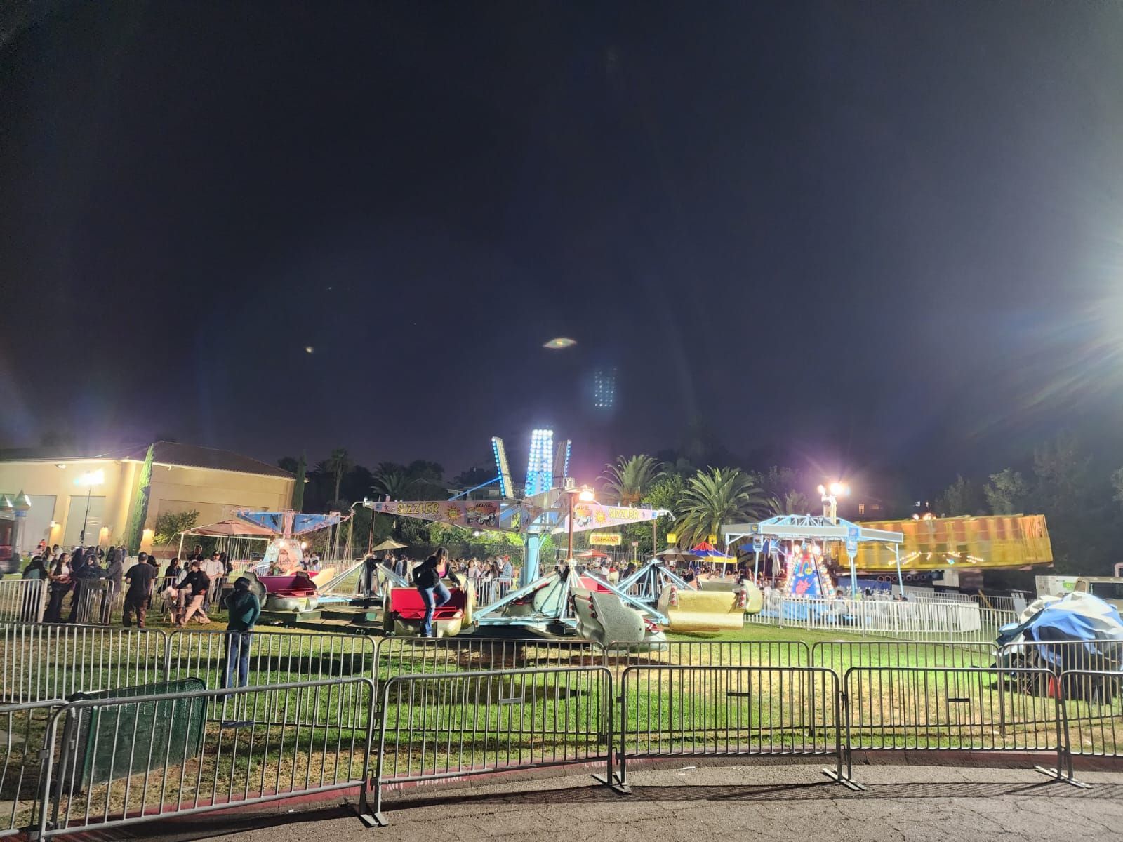 Nighttime fairground with bright rides, tents, and crowds behind a green fence.