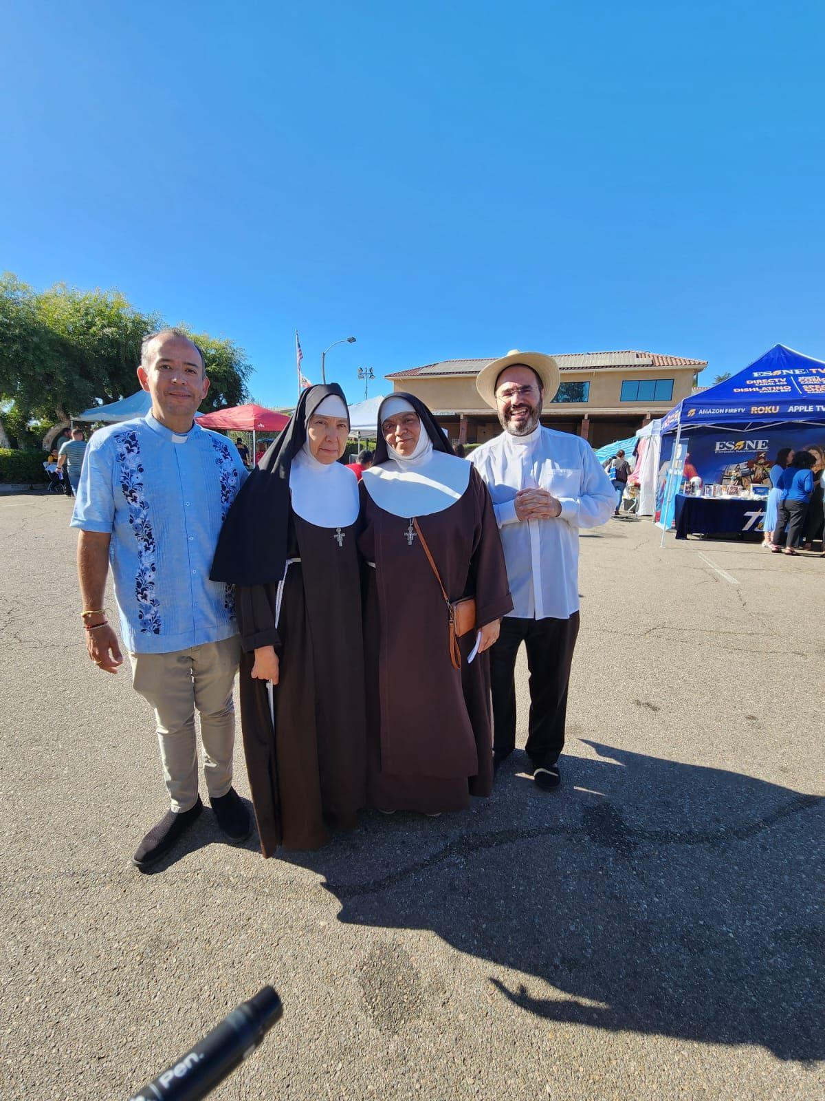 Four people posing outdoors on a sunny day, with blue sky and a tent in the background.