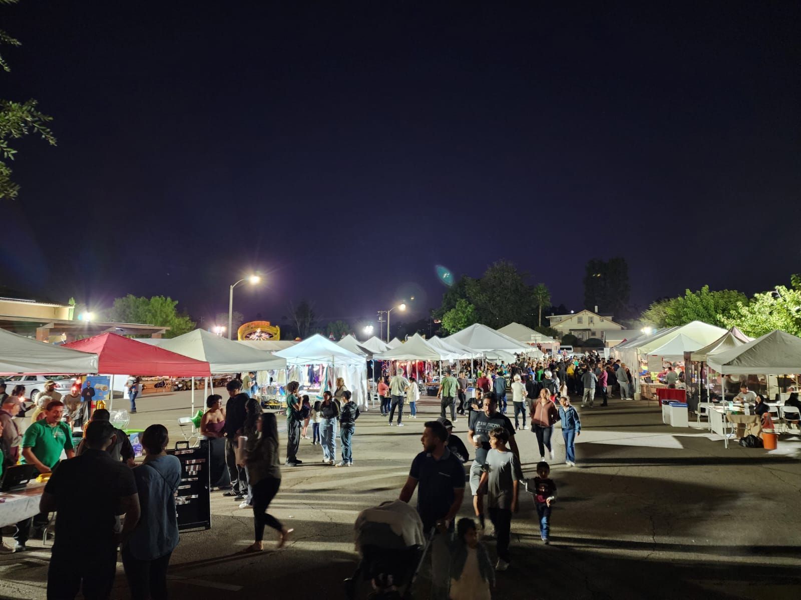 Nighttime outdoor market with white tents, bright lights, and a crowd walking through the fairground