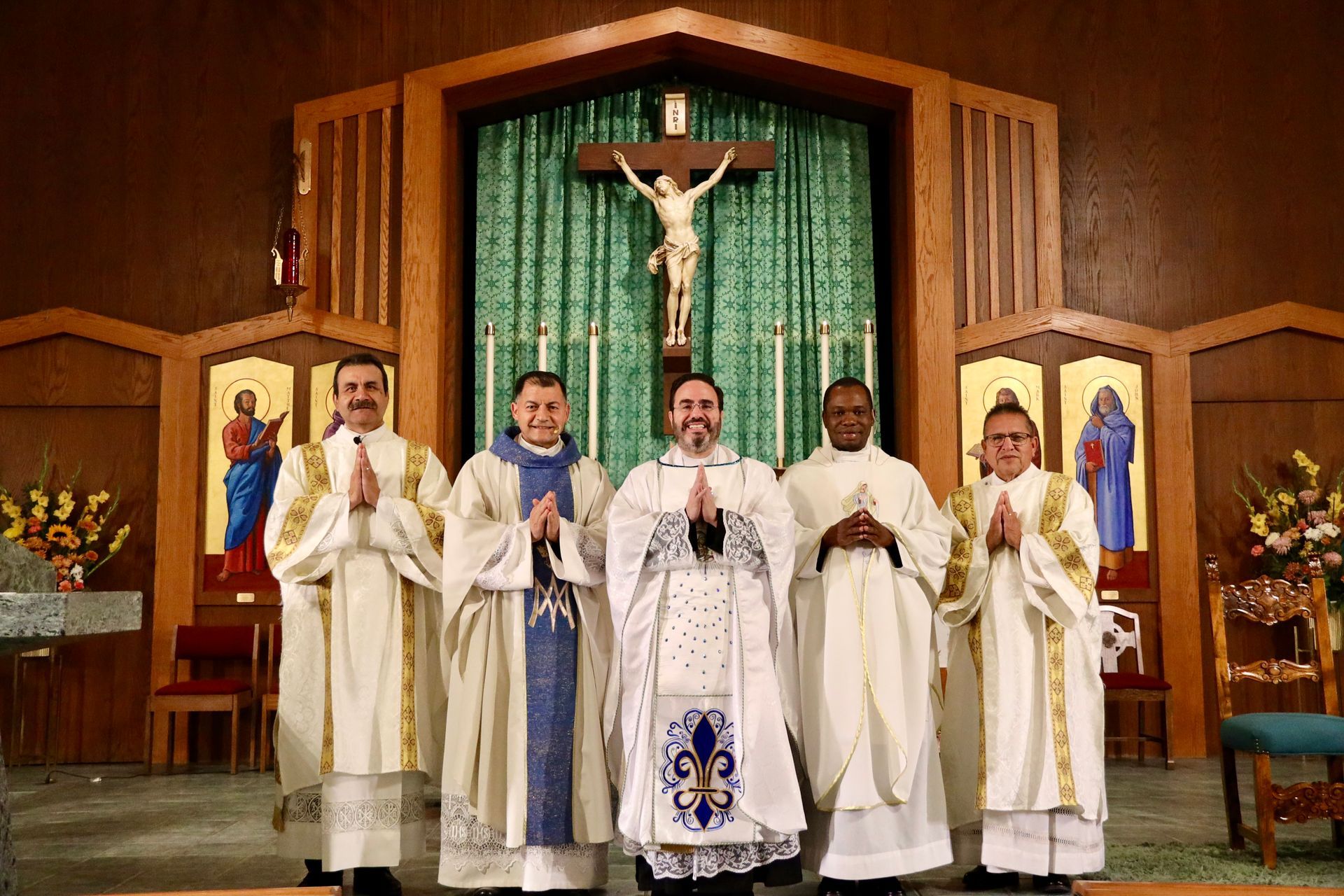 Six priests in white vestments stand before an altar with a crucifix in a church.