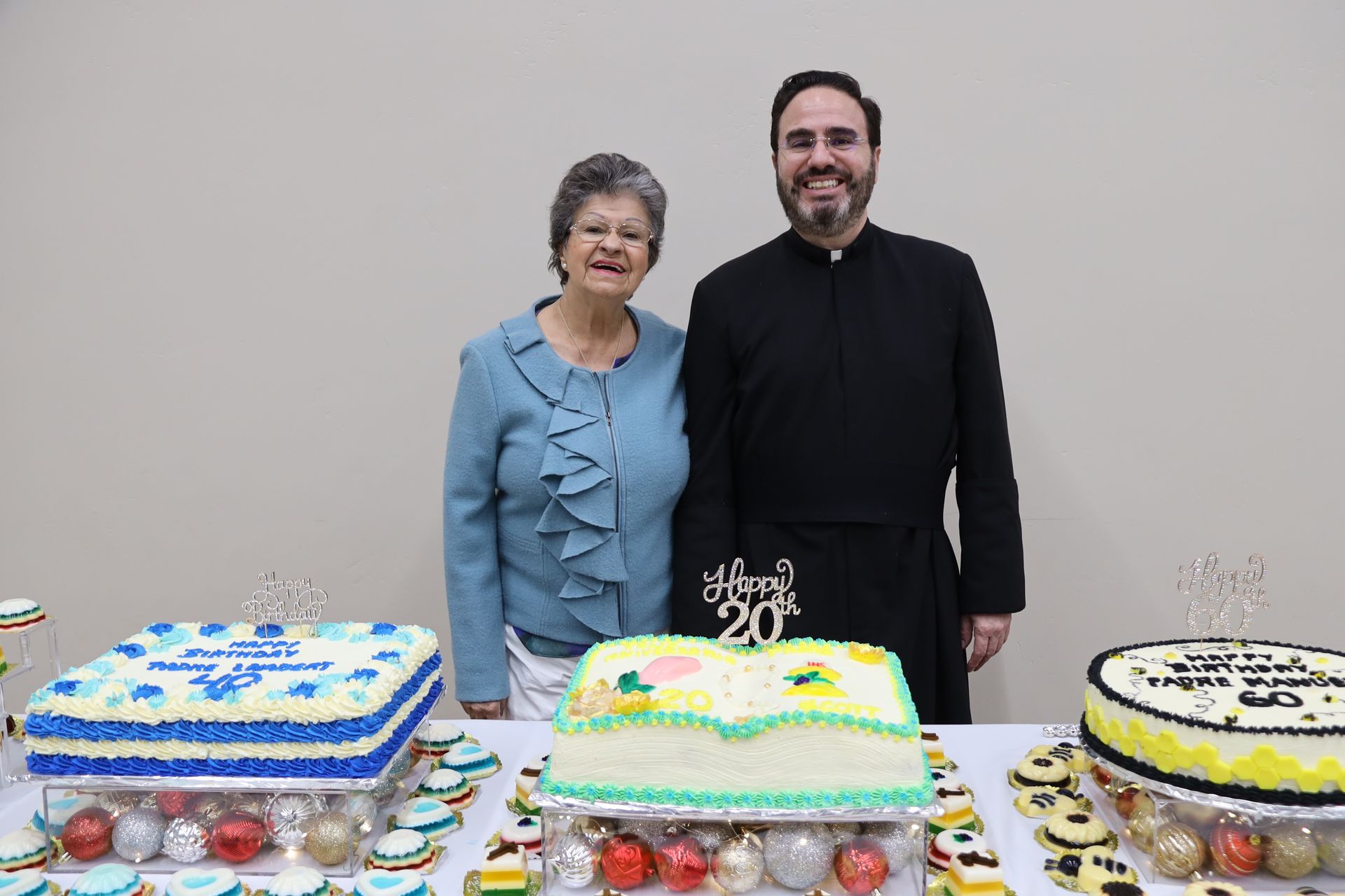 Two people standing behind three decorated cakes on a table, smiling at a celebration.