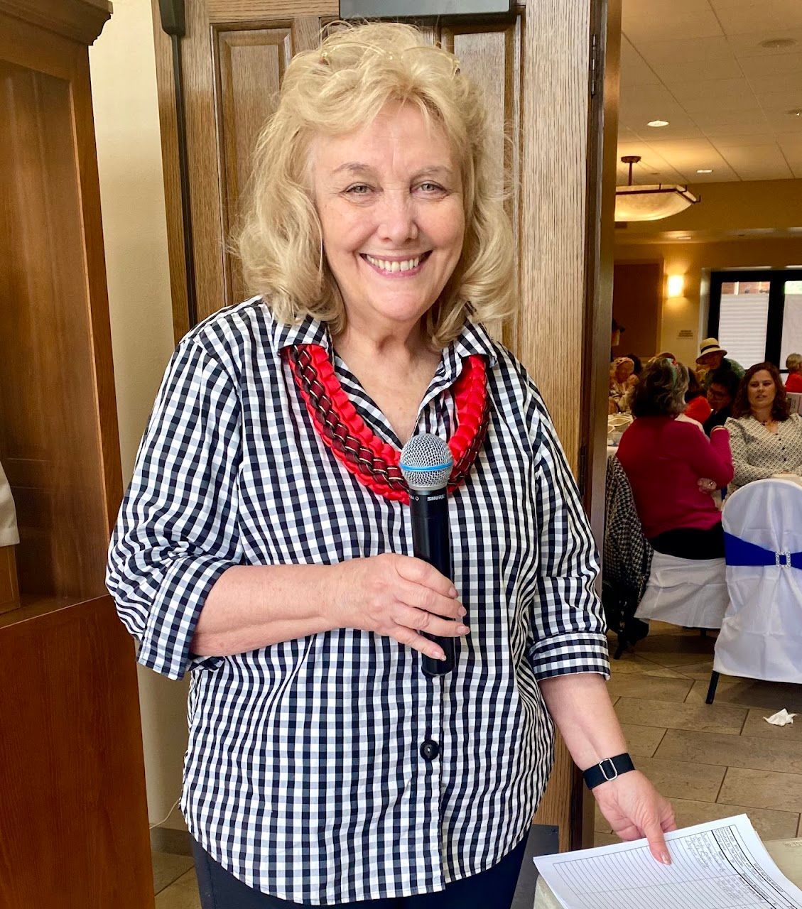 Smiling woman in a checkered shirt and red necklace stands indoors holding papers.