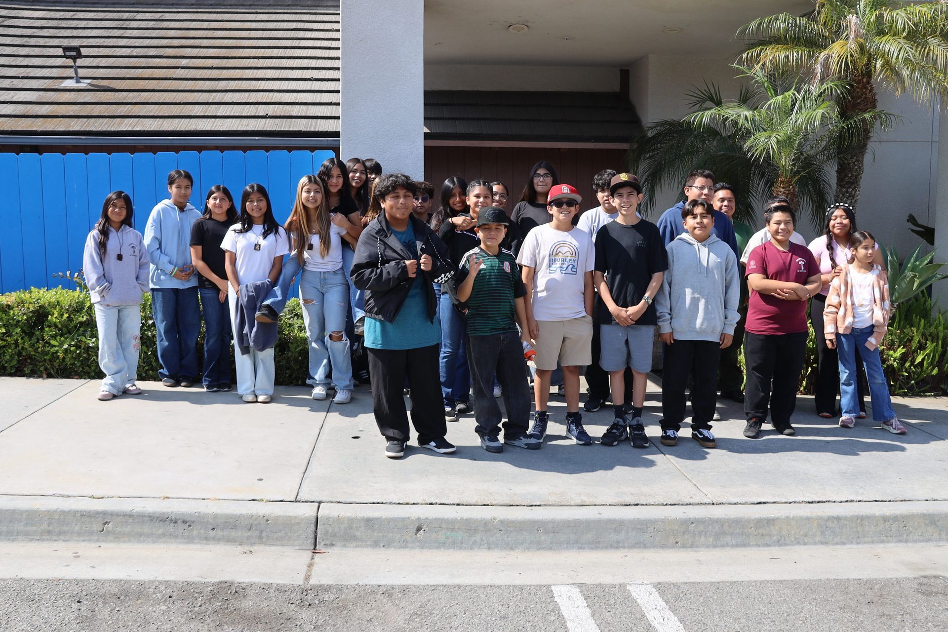 Group of people standing on a sidewalk outside a building, posing together in front of a blue wall.