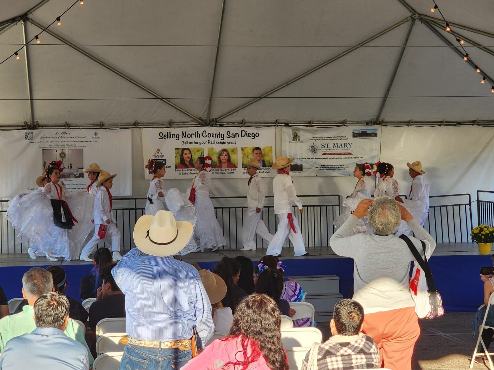 Costumed dancers perform on an outdoor stage under a tent before an audience