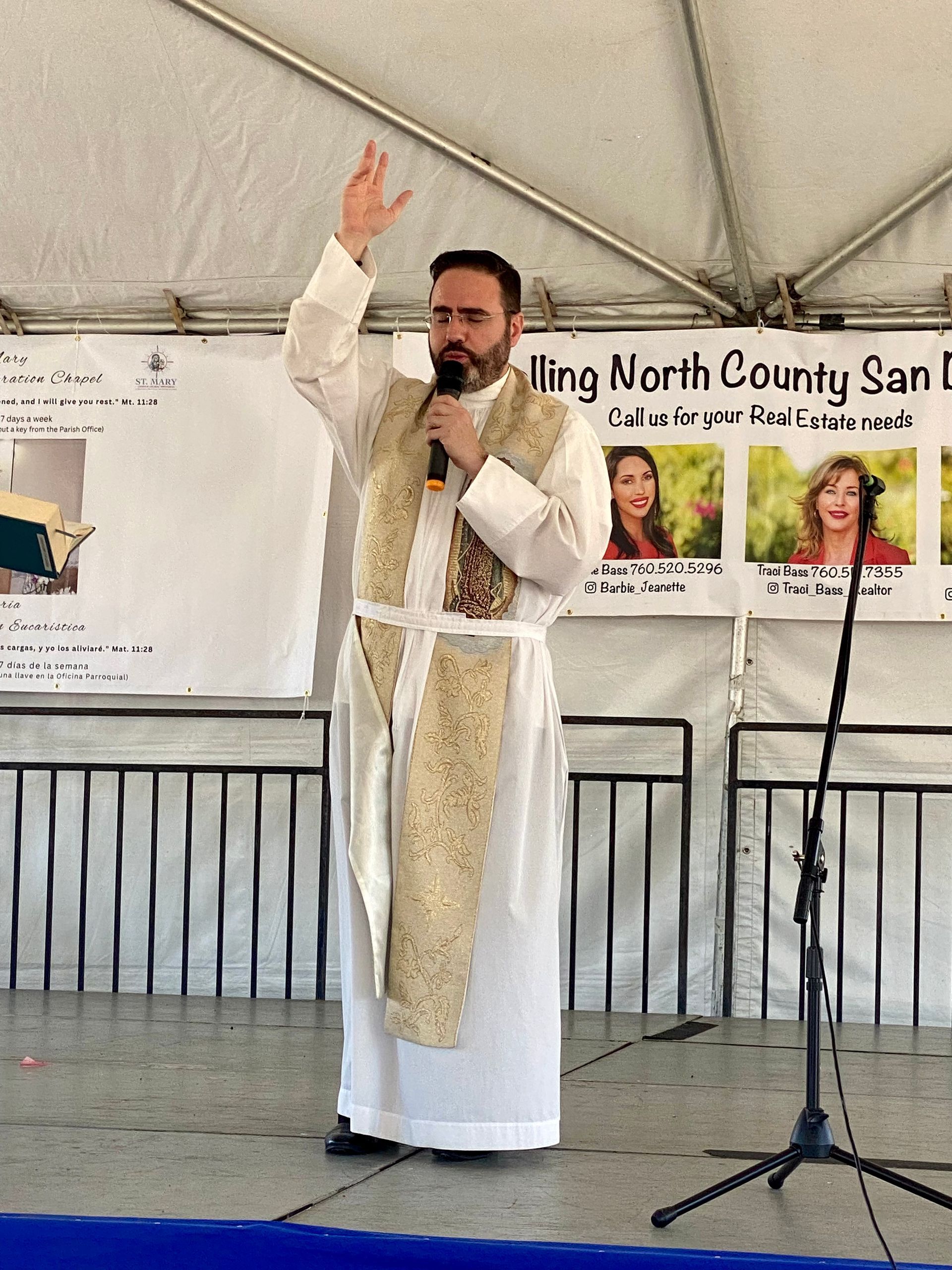 Man in white robe speaking on stage under a tent, holding a microphone and raising one hand.